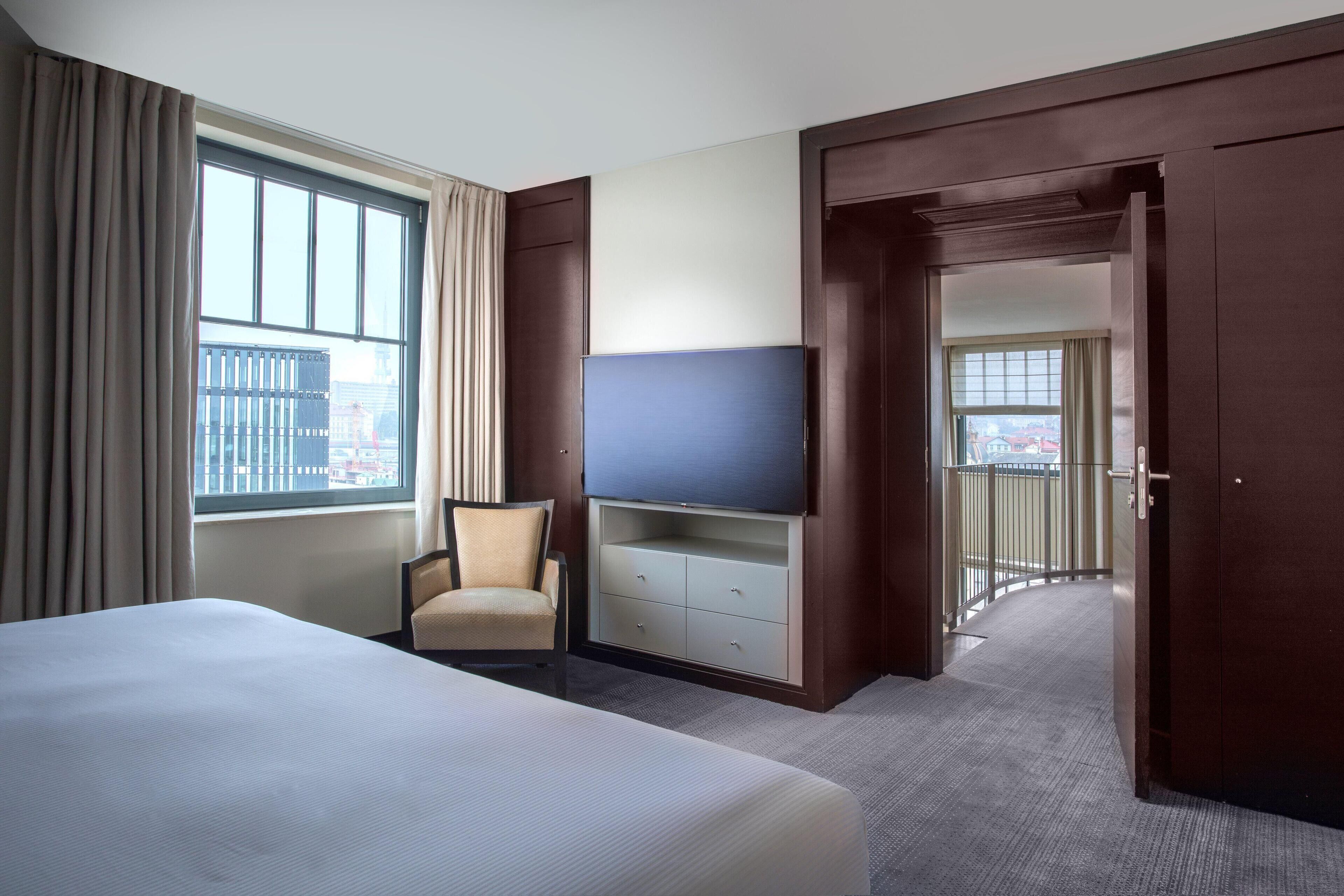 Habitación de hotel con una cama grande, un televisor sobre un mueble de madera, una silla y vistas a la ciudad a través de la ventana.