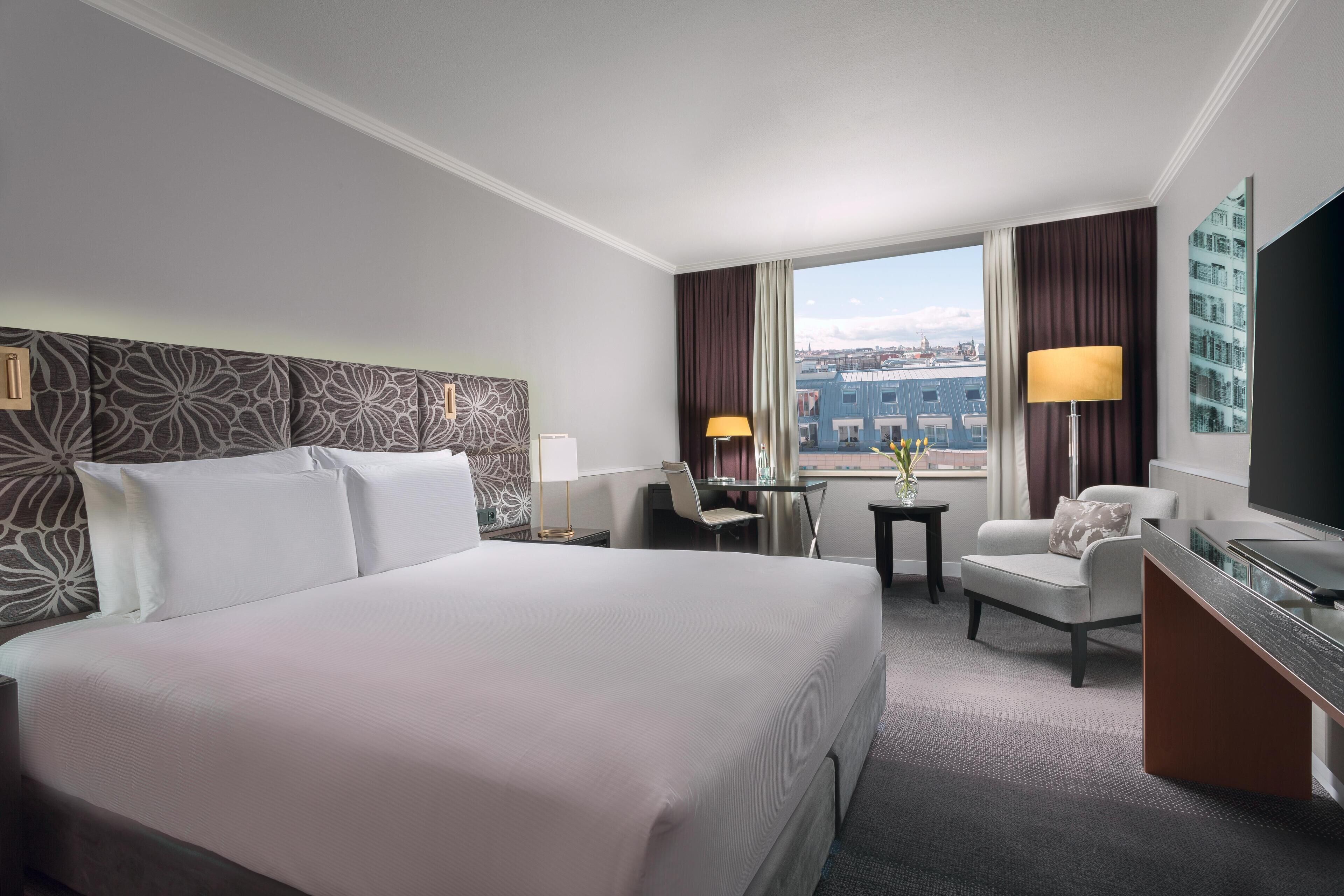 A hotel room with a large bed, a desk, a chair, a TV, and a view of the city through the window.