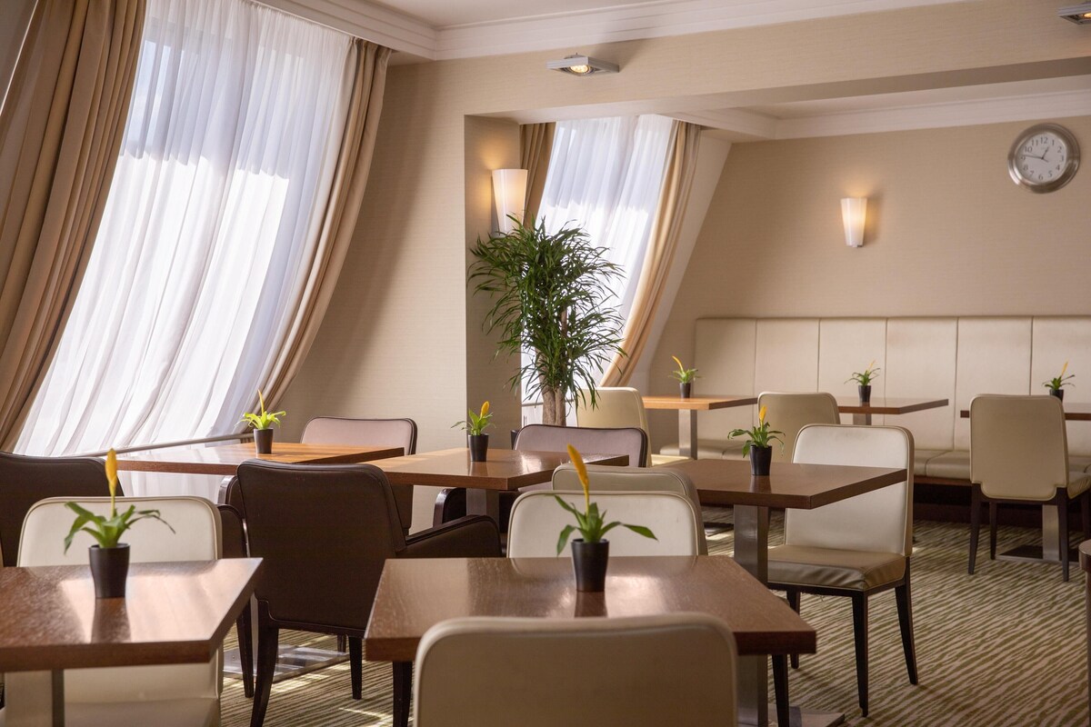 A hotel room with a dining area featuring tables and chairs, a clock on the wall, and potted plants.