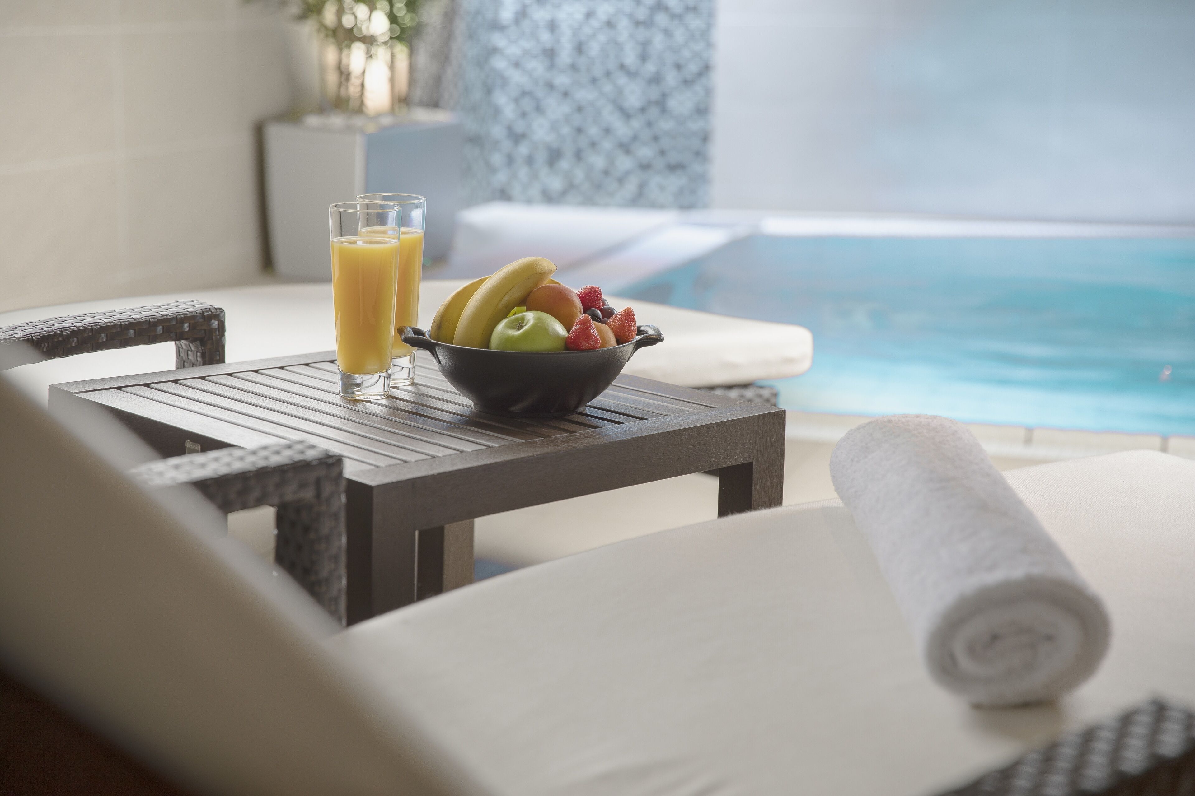 Ett matbord med en skål med frukt och ett glas apelsinjuice, en hoprullad handduk och utsikt över en pool.