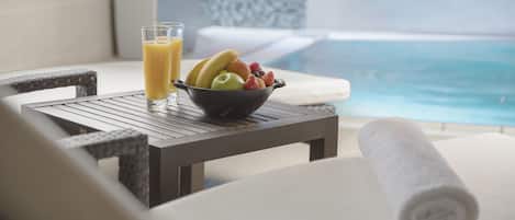 A dining table with a bowl of fruit and a glass of orange juice, a rolled towel, and a view of a pool.
