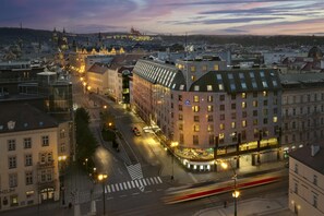 A hotel with a modern glass facade, located in a city center with historic buildings in the background.