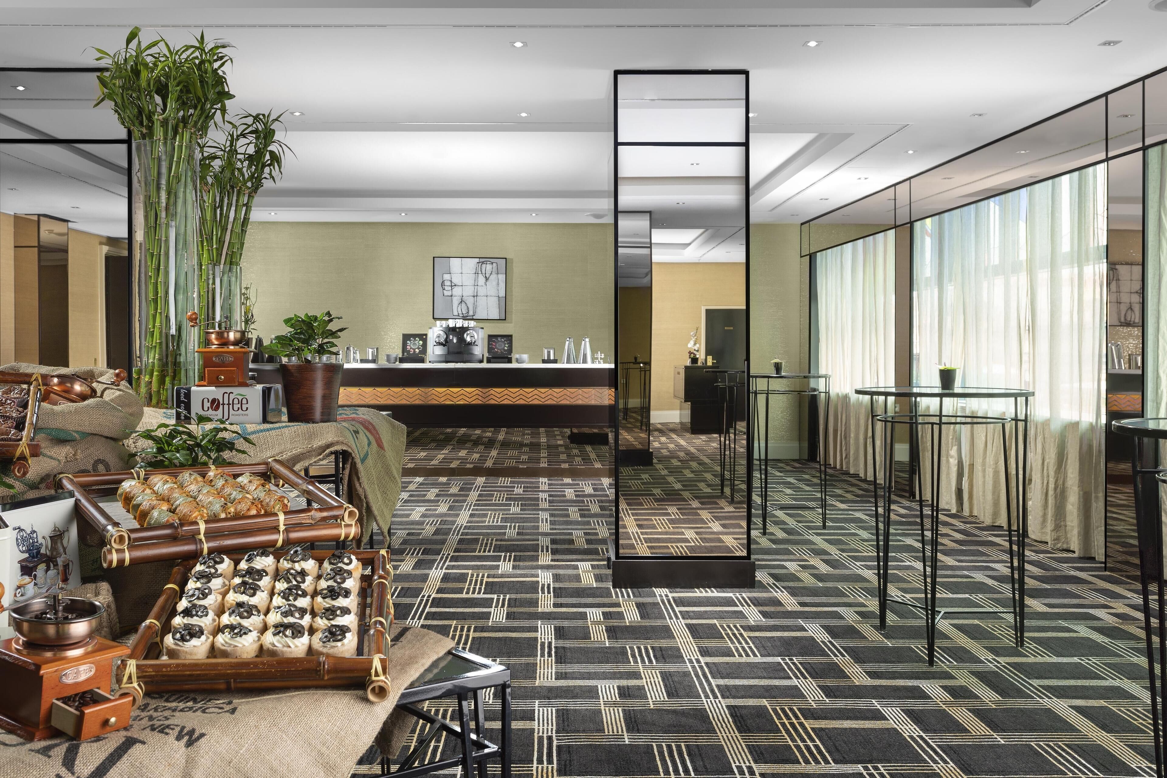 A modern hotel lobby with a coffee station, bamboo plants, and a patterned carpet.