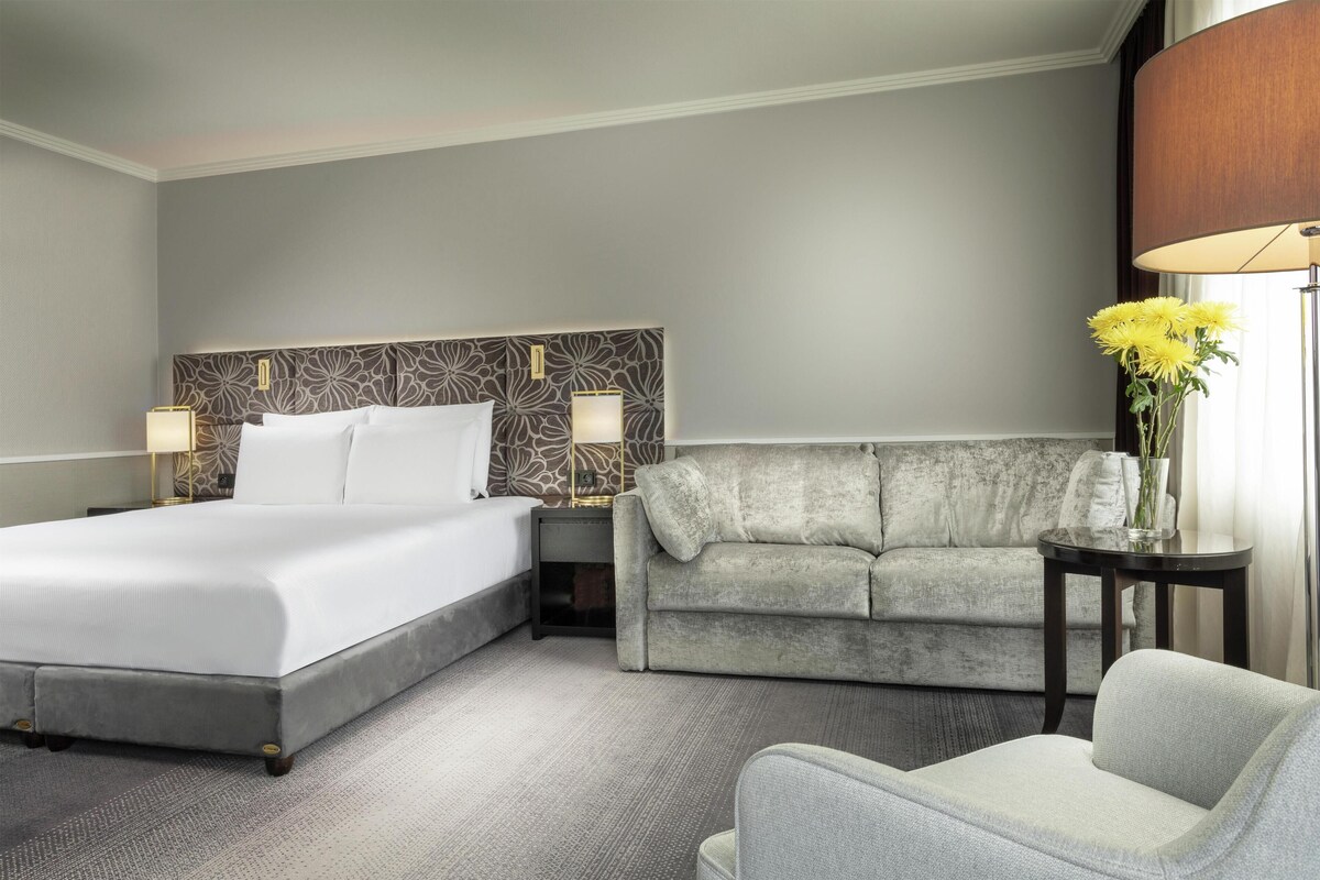 A hotel room with a large bed, a grey sofa, a small table with a vase of flowers, and a lamp.