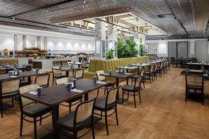 A spacious dining area with multiple tables and chairs, a central seating area with a green sofa, and a kitchen in the background. - Hilton Prague Old Town (Prague)