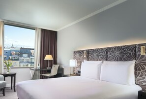 A hotel room with a large bed, a desk, a chair, and a view of the cityscape.
