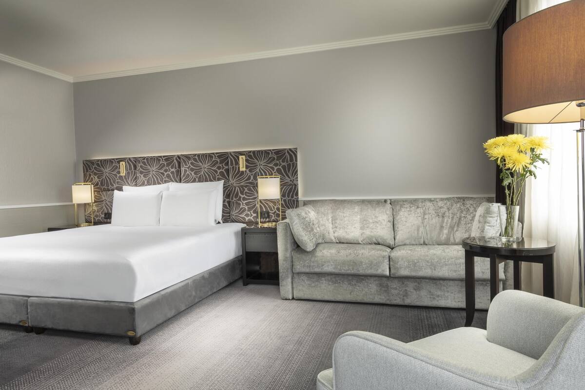 Habitación de hotel con una cama grande, un sofá gris, una mesita con un florero y una lámpara.