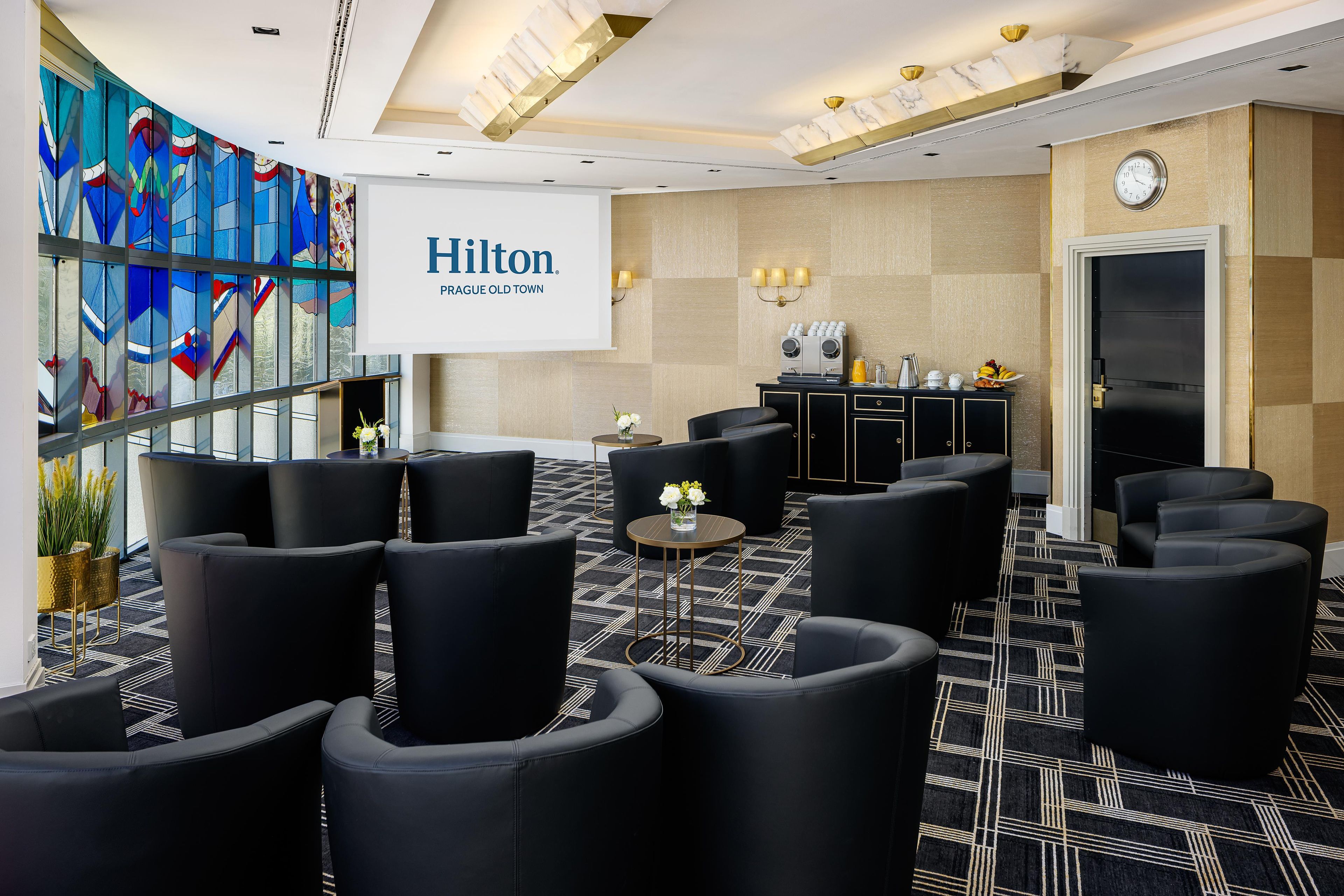 Um saguão de hotel moderno com poltronas de couro preto, um espaço para café e uma tela grande exibindo o nome do hotel.