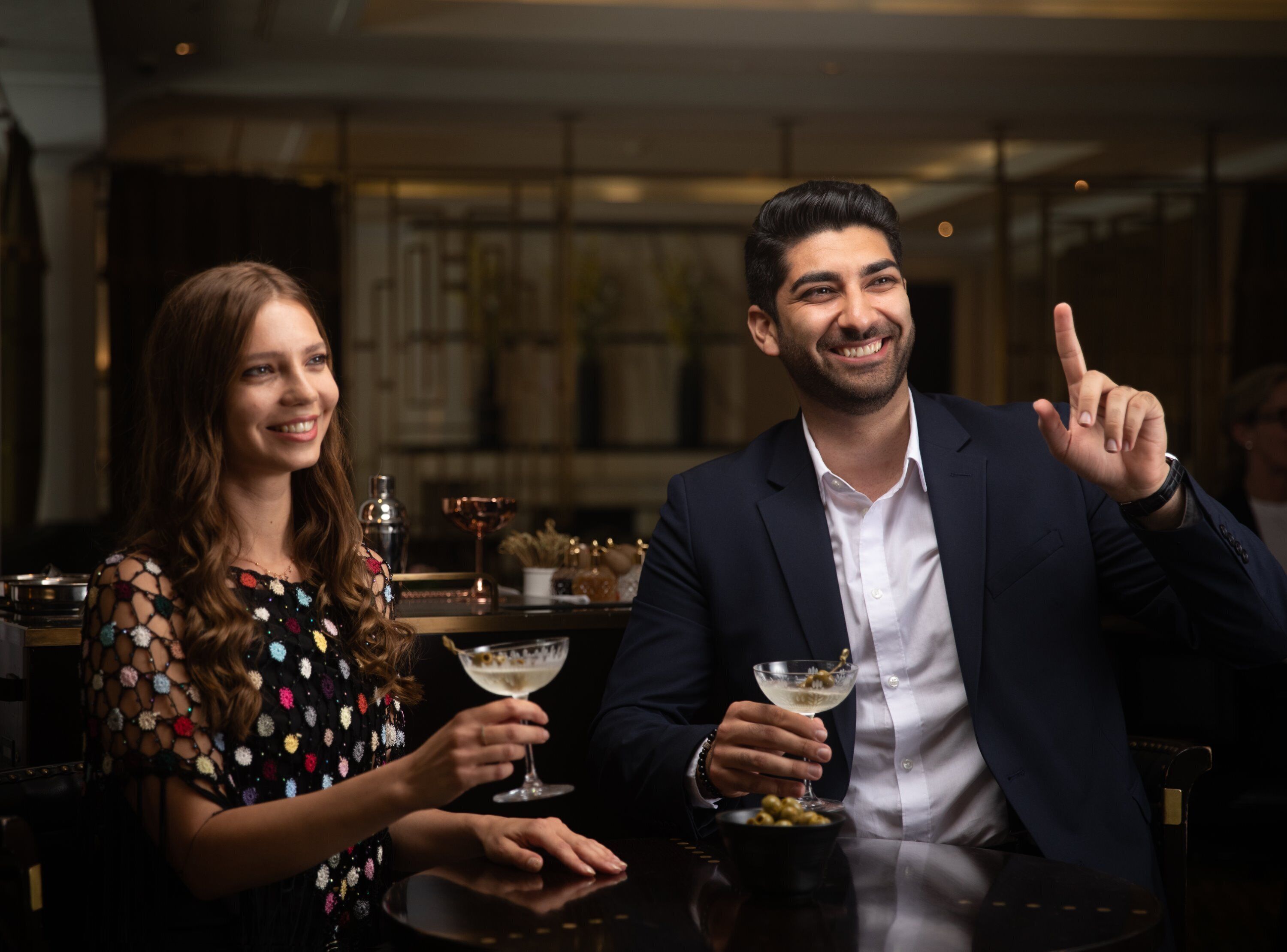 Um homem e uma mulher sentados à mesa num espaço com pouca luz, com um bar ao fundo.