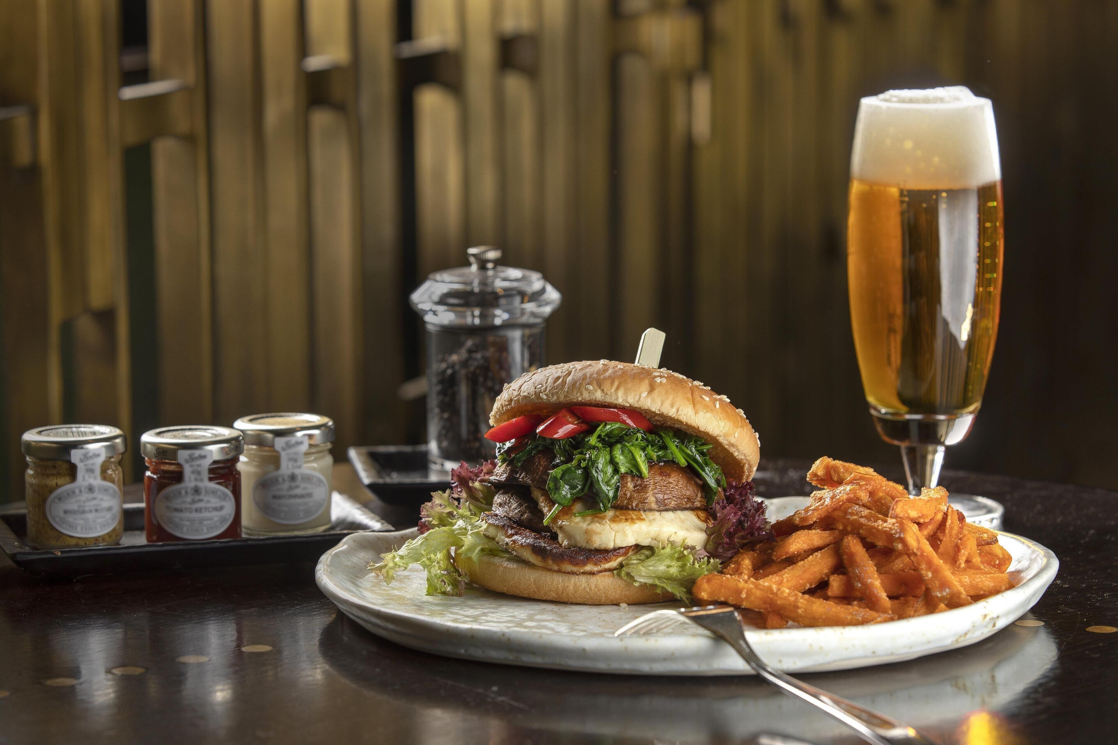 Uma refeição com um hambúrguer, batatas fritas e um copo de cerveja sobre uma mesa.