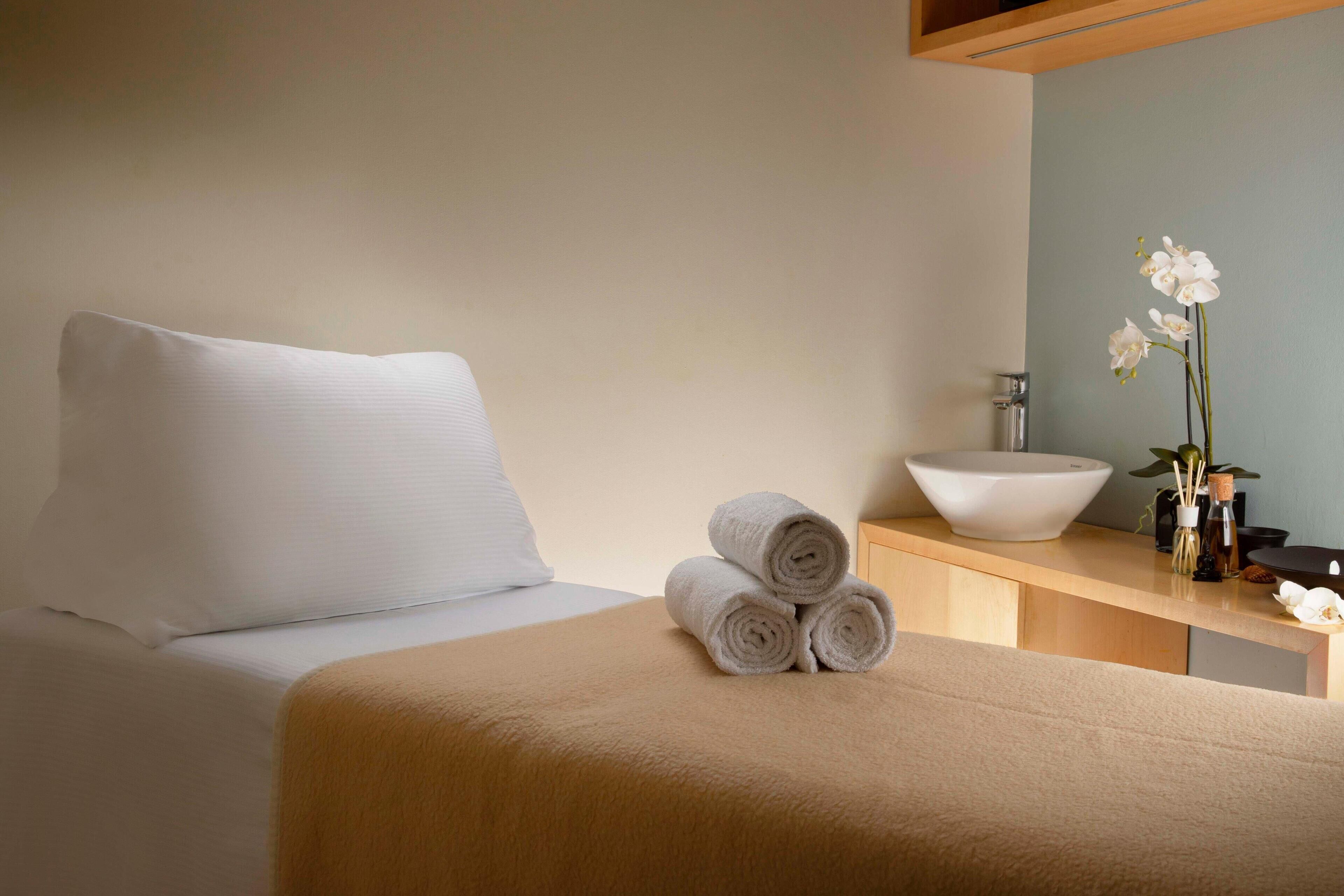 Quarto de hotel com uma cama, uma mesa de cabeceira de madeira, uma tigela branca e um vaso com flores.