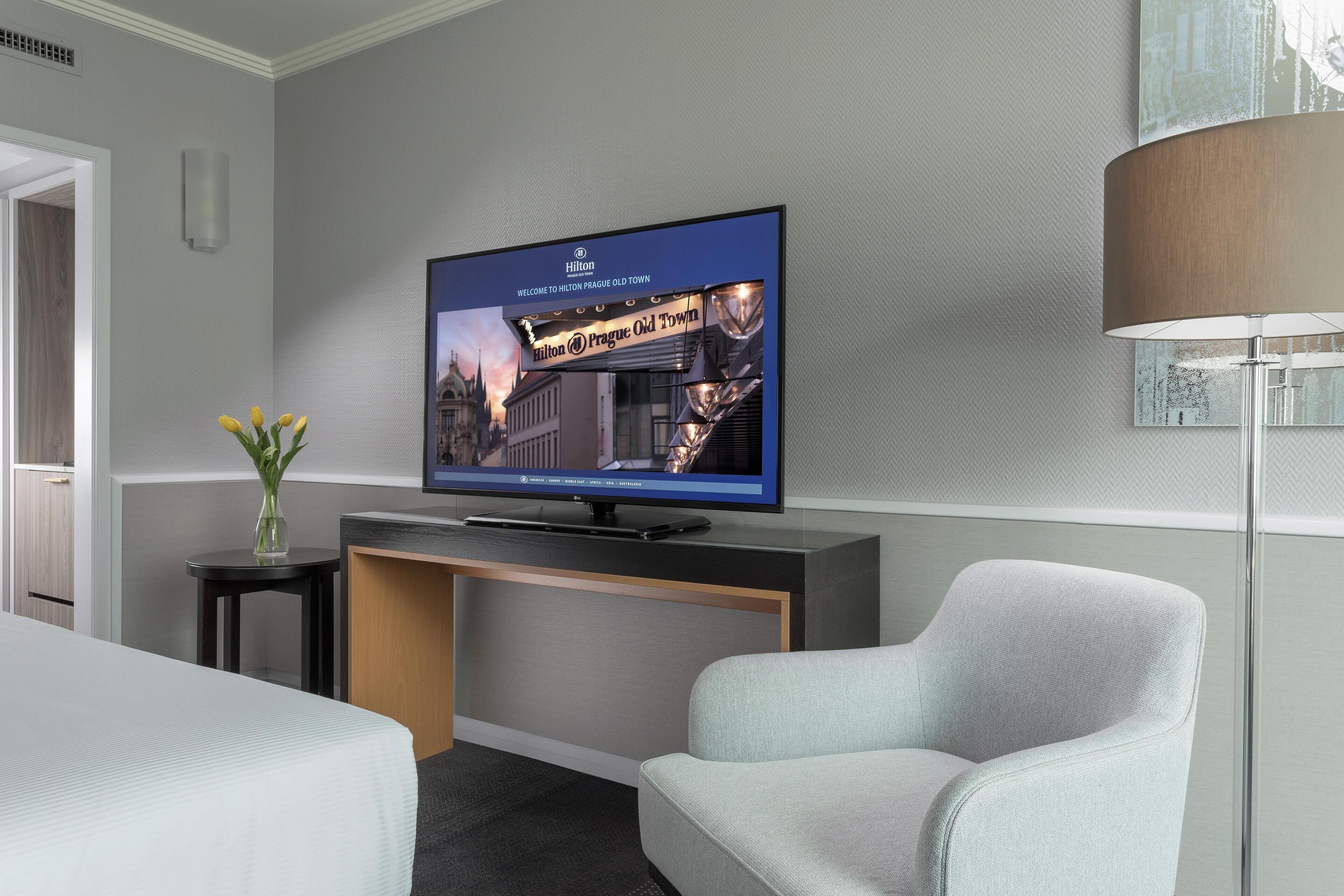 Una camera d'albergo moderna con una televisione a schermo piatto che mostra una vista sulla città, una comoda poltrona grigio chiaro, un tavolino con un vaso di fiori e una scrivania con una lampada.