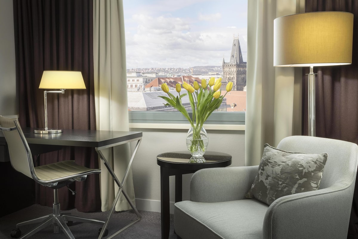 Habitación de hotel con escritorio, silla, lámpara y vistas a un paisaje urbano a través de una ventana.