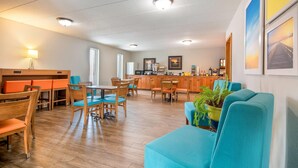 A hotel lobby with wooden flooring, a dining area with orange and blue chairs, a kitchen area, and a seating area with a blue sofa and a potted plant. - Days Inn by Wyndham International Falls (International Falls)