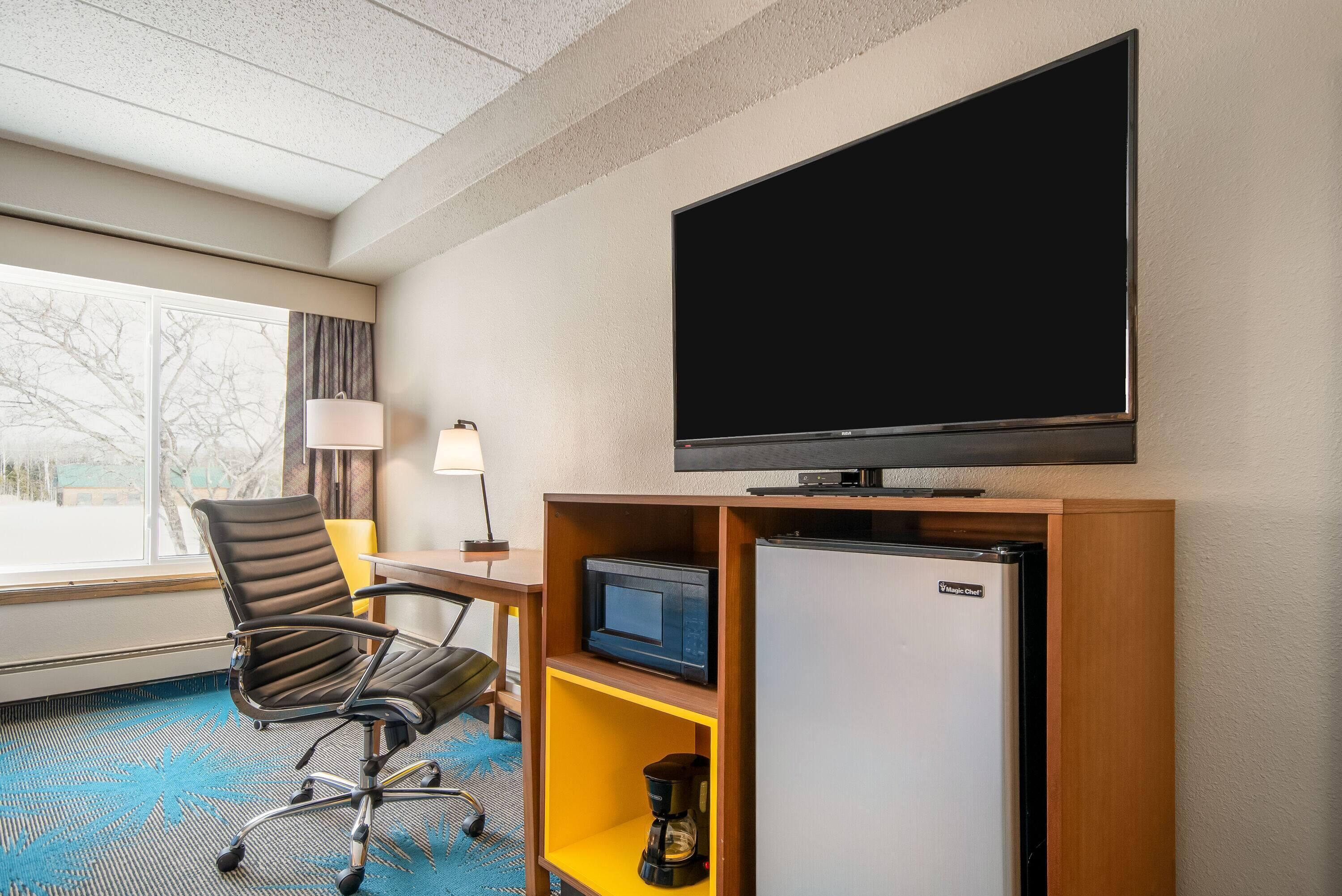 A hotel room with a large flat-screen TV, a microwave, a refrigerator, a coffee maker, a desk, a chair, and a window with a view of trees.