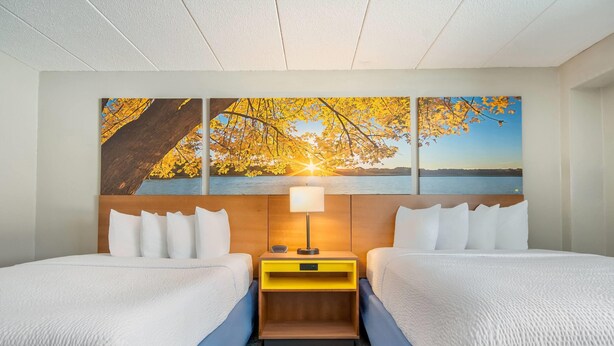 A hotel room with two beds, a wooden headboard, a nightstand with a lamp, and a large painting of a sunset over a lake. - Days Inn by Wyndham International Falls (International Falls)
