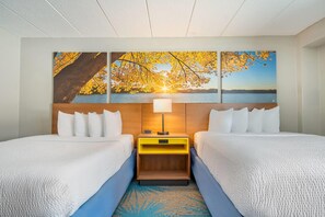 A hotel room with two beds, a wooden headboard, a nightstand with a lamp, and a large painting of a sunset over a lake.
