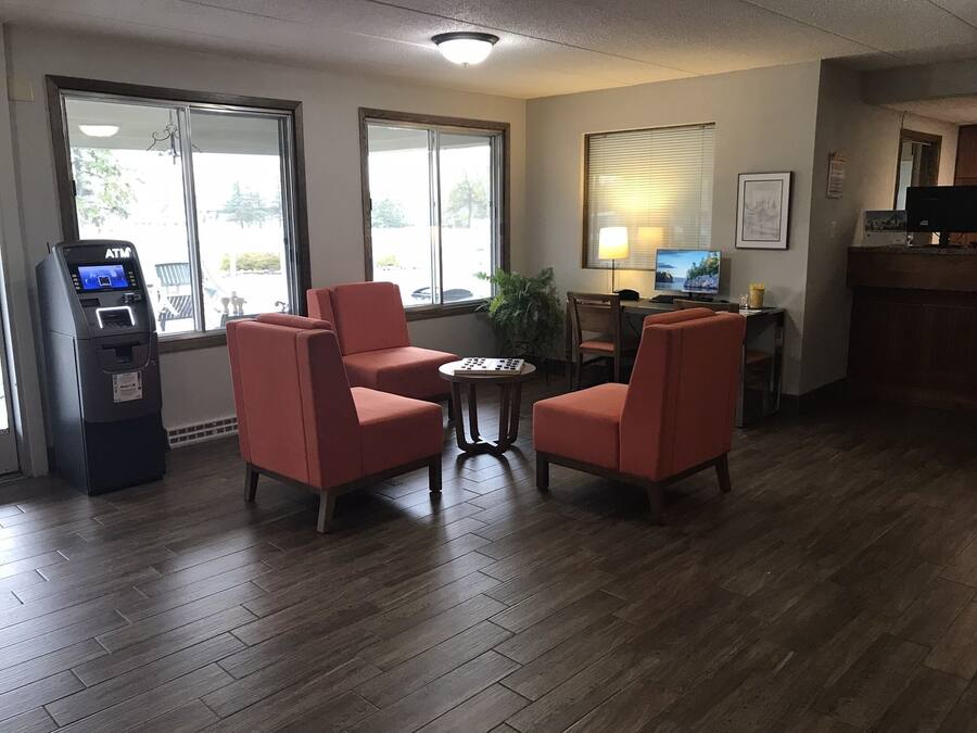 A hotel lobby with wooden flooring, red armchairs, a small table, an ATM, a TV, and a window with blinds.