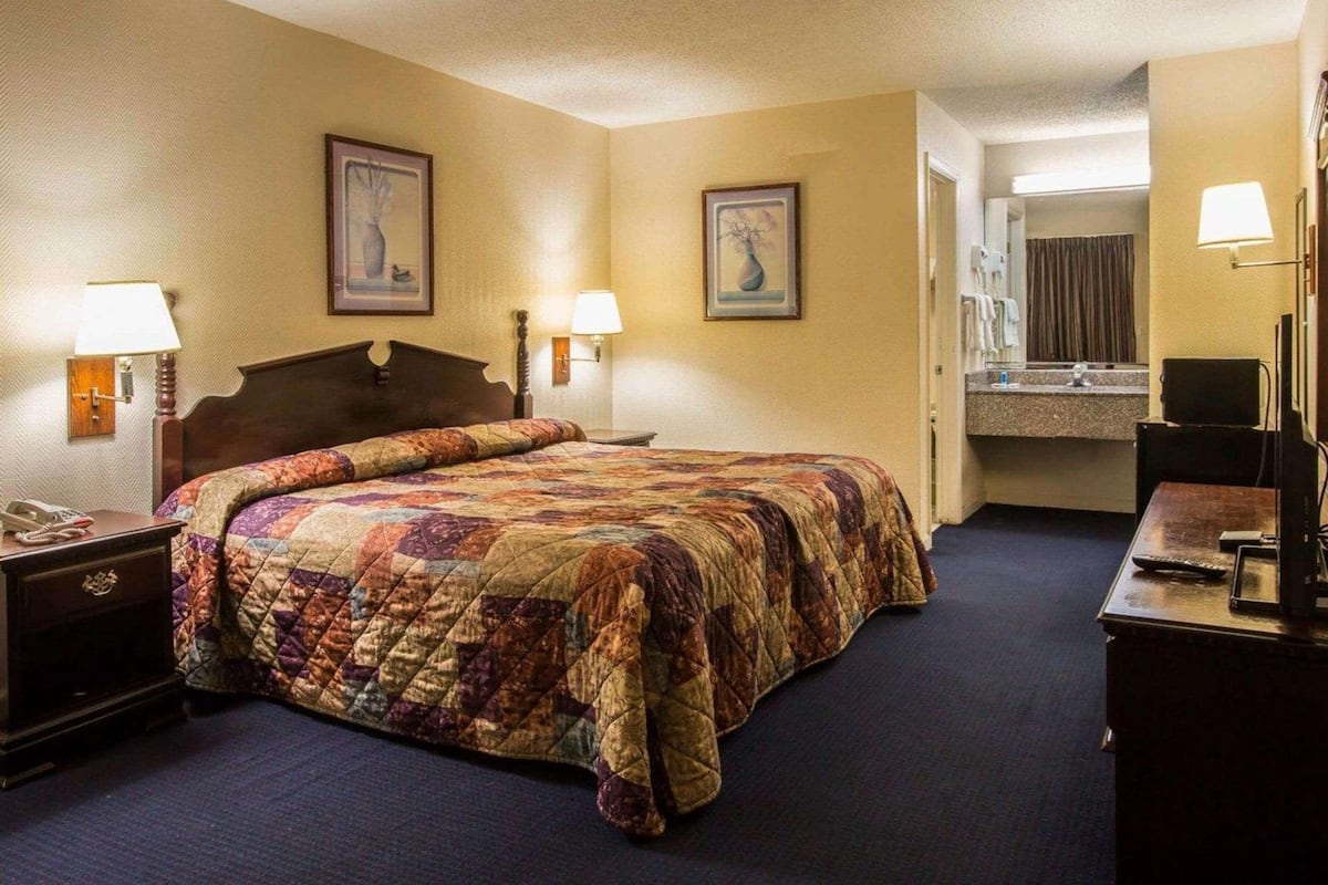 Une chambre d’hôtel avec un grand lit, deux lampes de chevet, une table de chevet, une commode et une salle de bain visible à travers une porte ouverte.