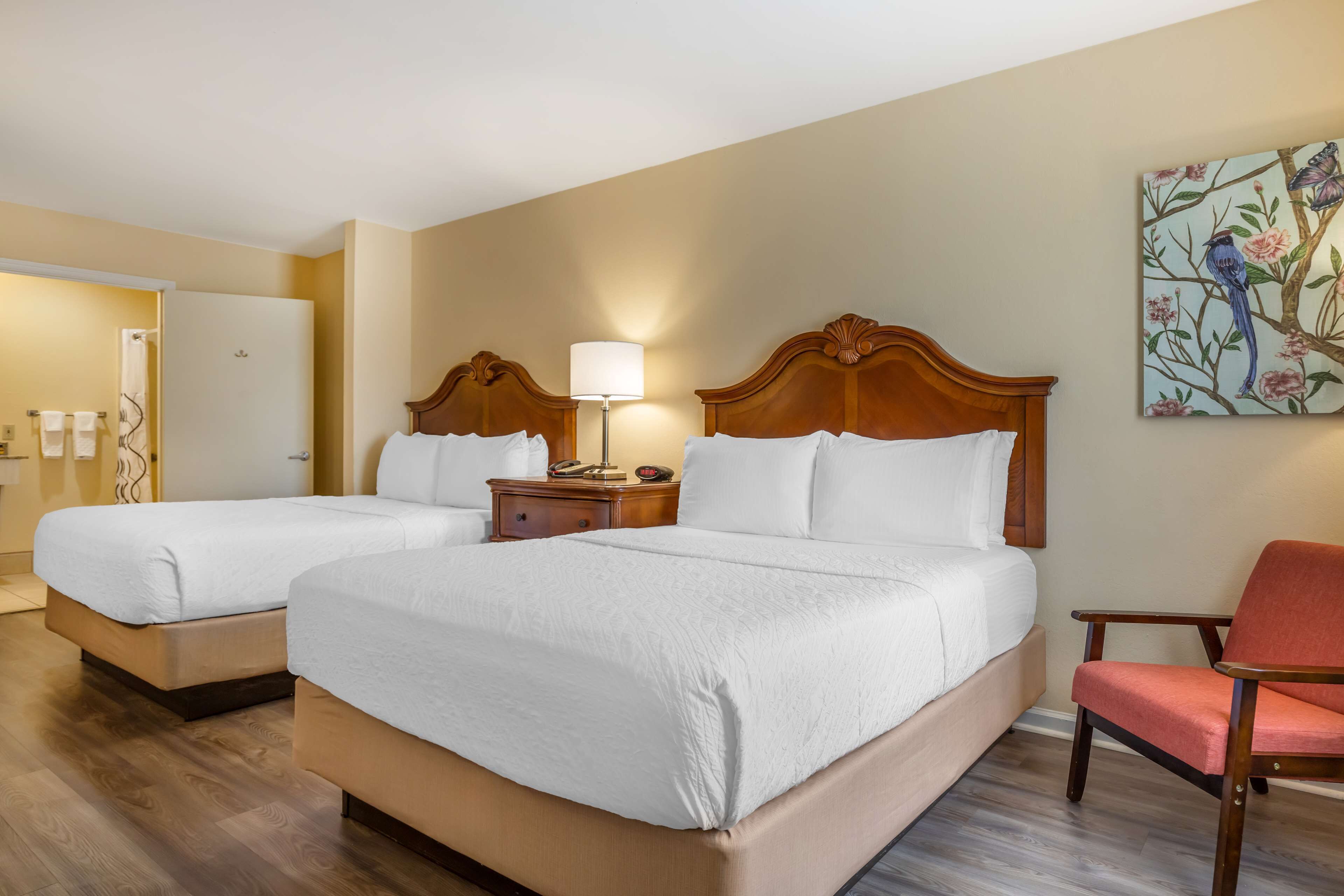 Una habitación de hotel con dos camas individuales, un cabecero de madera, una mesita de noche con lámpara, una silla roja y una obra de arte floral en la pared.