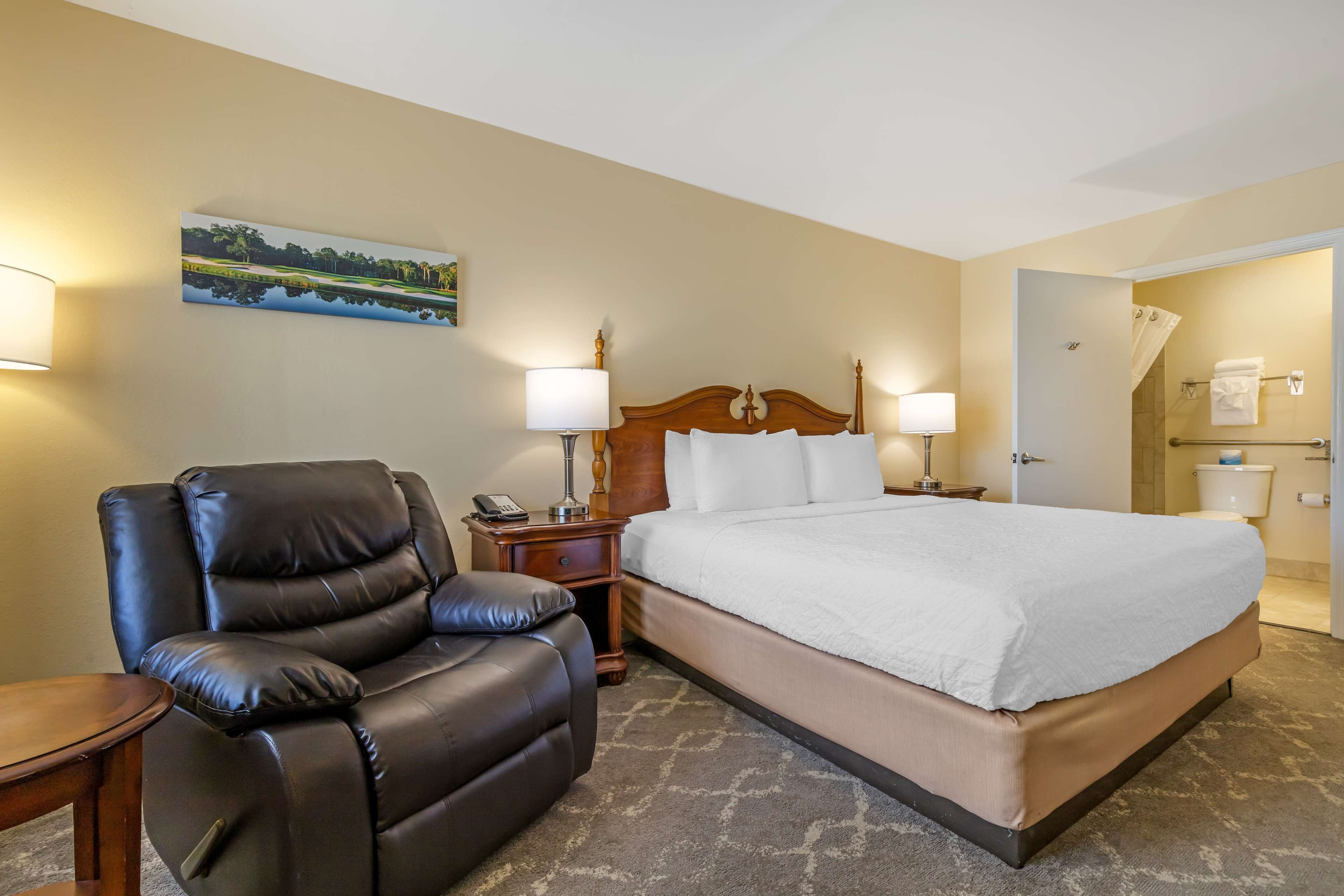 Habitación de hotel con una cama grande, un sillón de cuero, lámparas de noche, un cuadro en la pared y una puerta de baño.