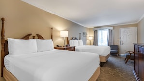 A hotel room with two large beds, a wooden headboard, bedside tables with lamps, a wardrobe, and a view of the outdoors through a window.