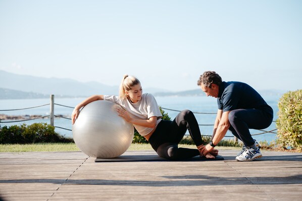 Dos personas ejercitan con una pelota grande de gimnasia sobre una cubierta de madera cerca de agua.