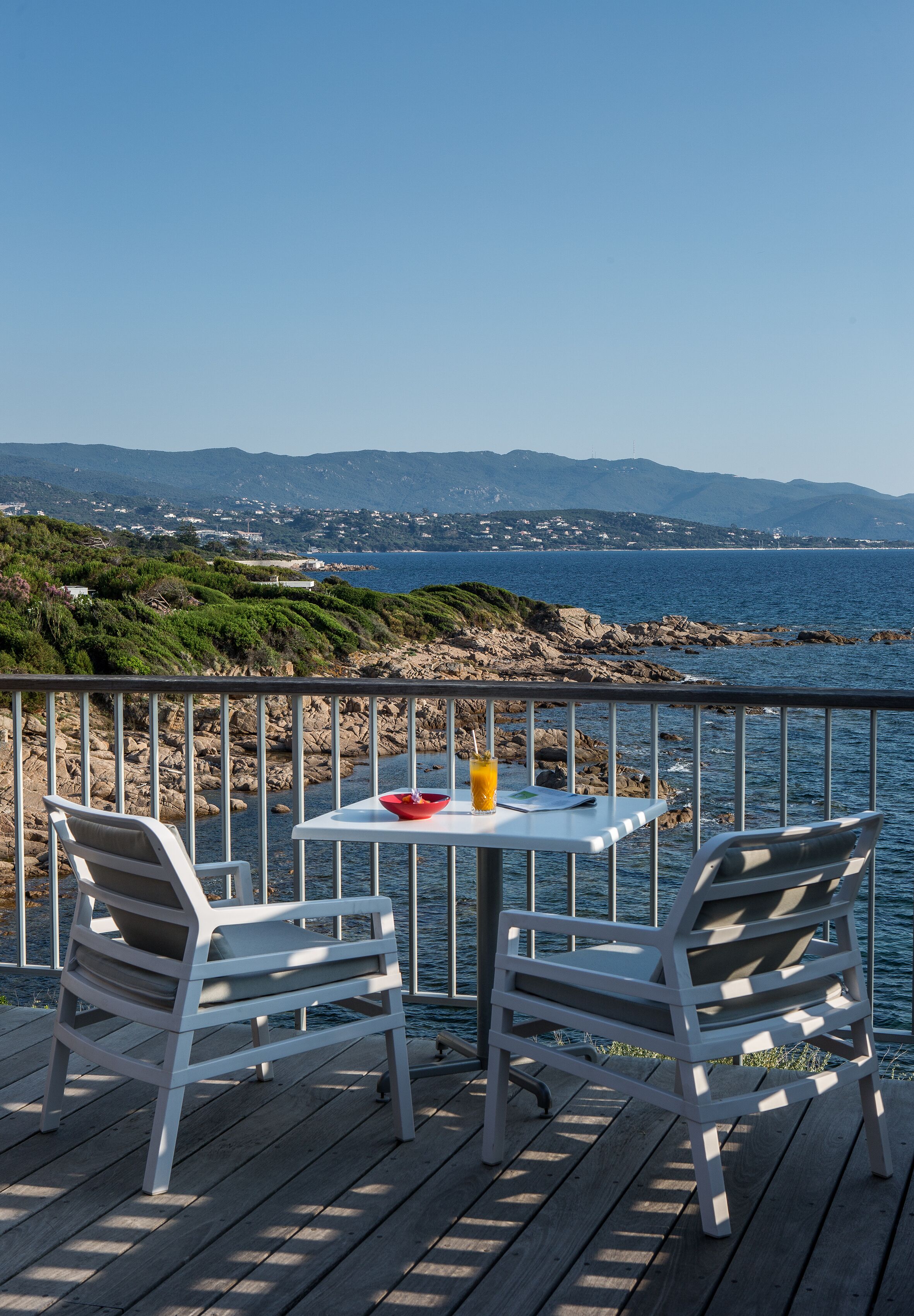 A deck with two white chairs and a small table, overlooking a rocky coastline and the sea.