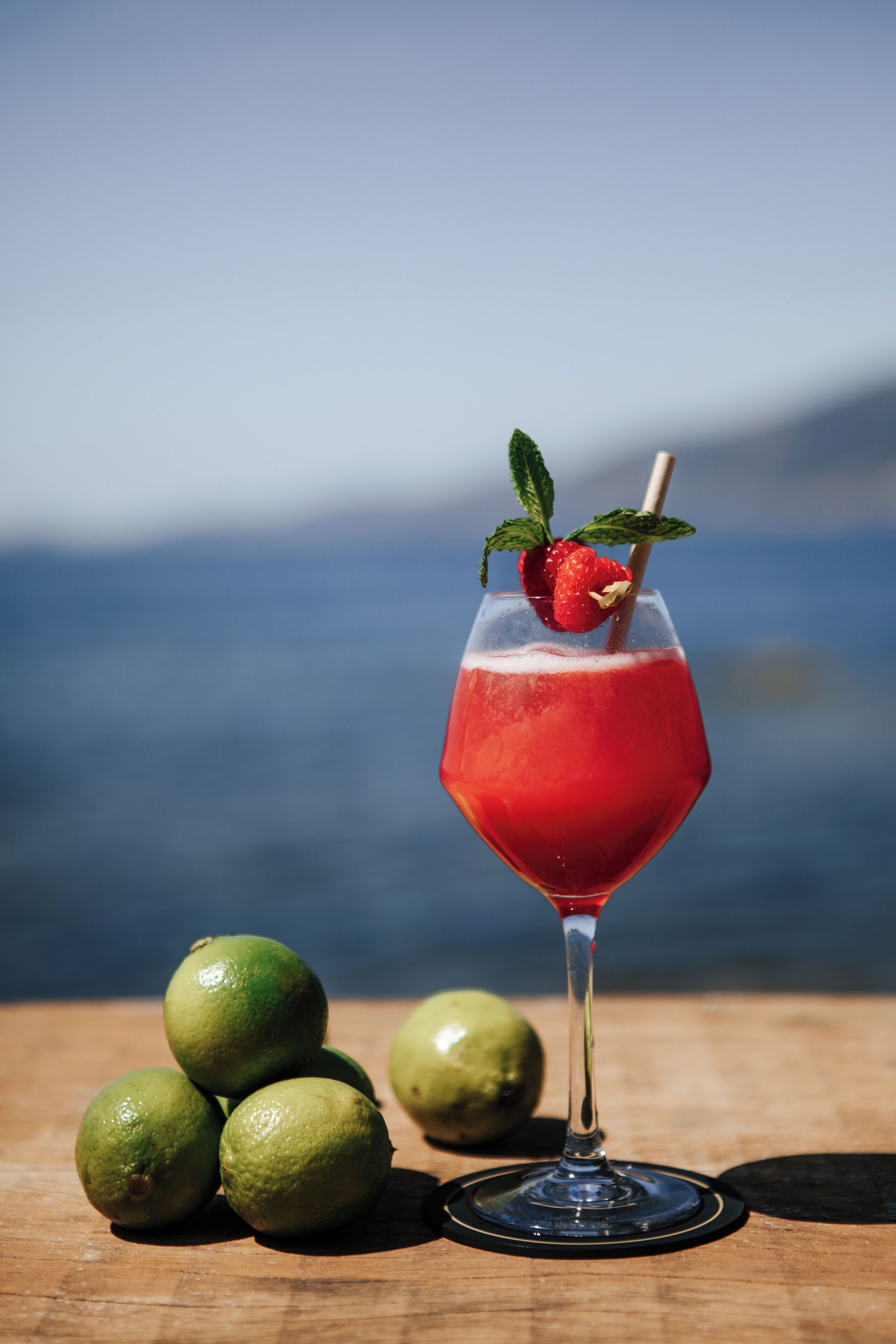 A cocktail glass with a red drink, garnished with a strawberry and mint, on a wooden table with limes.