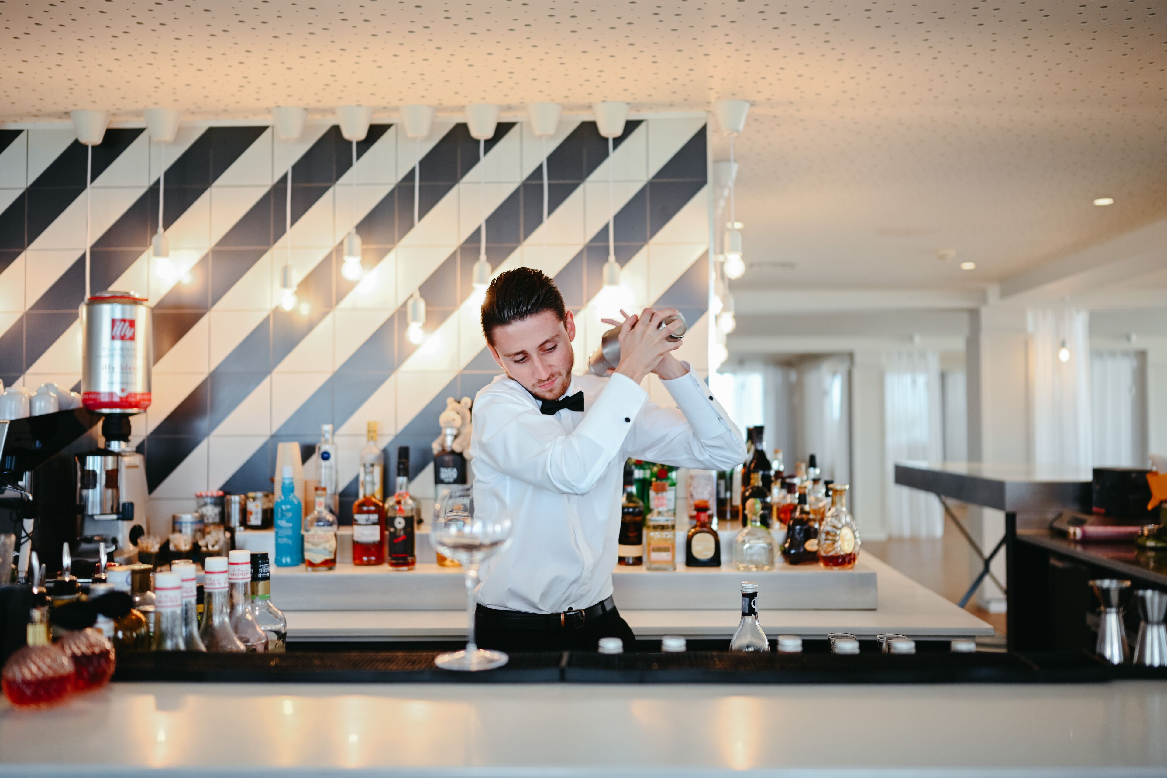 A bartender preparing a drink at a bar with a modern geometric ceiling design and a variety of bottles and glasses on the counter.