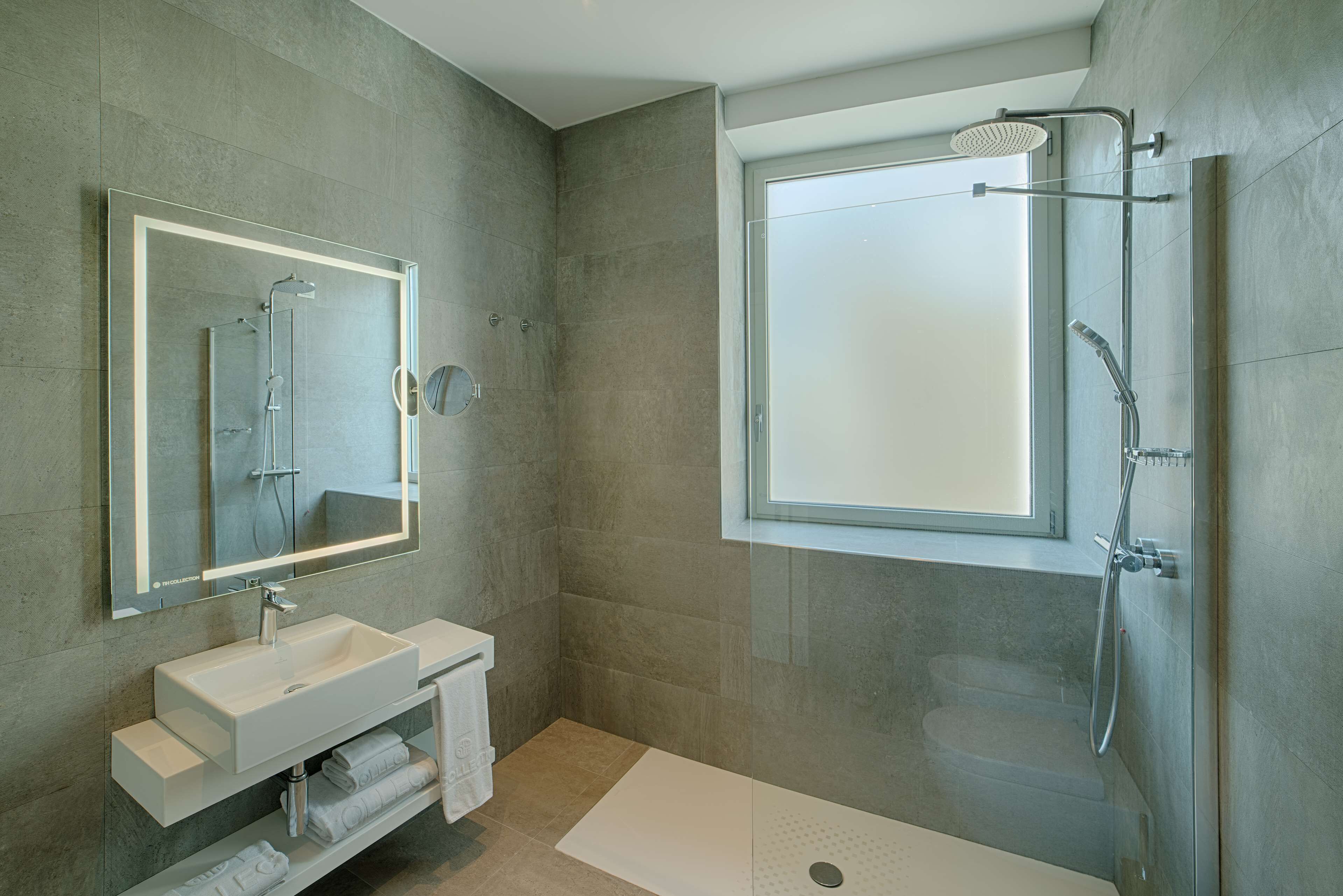 Un baño moderno con un espejo grande, un lavabo blanco, una ducha a ras de suelo y una ventana.