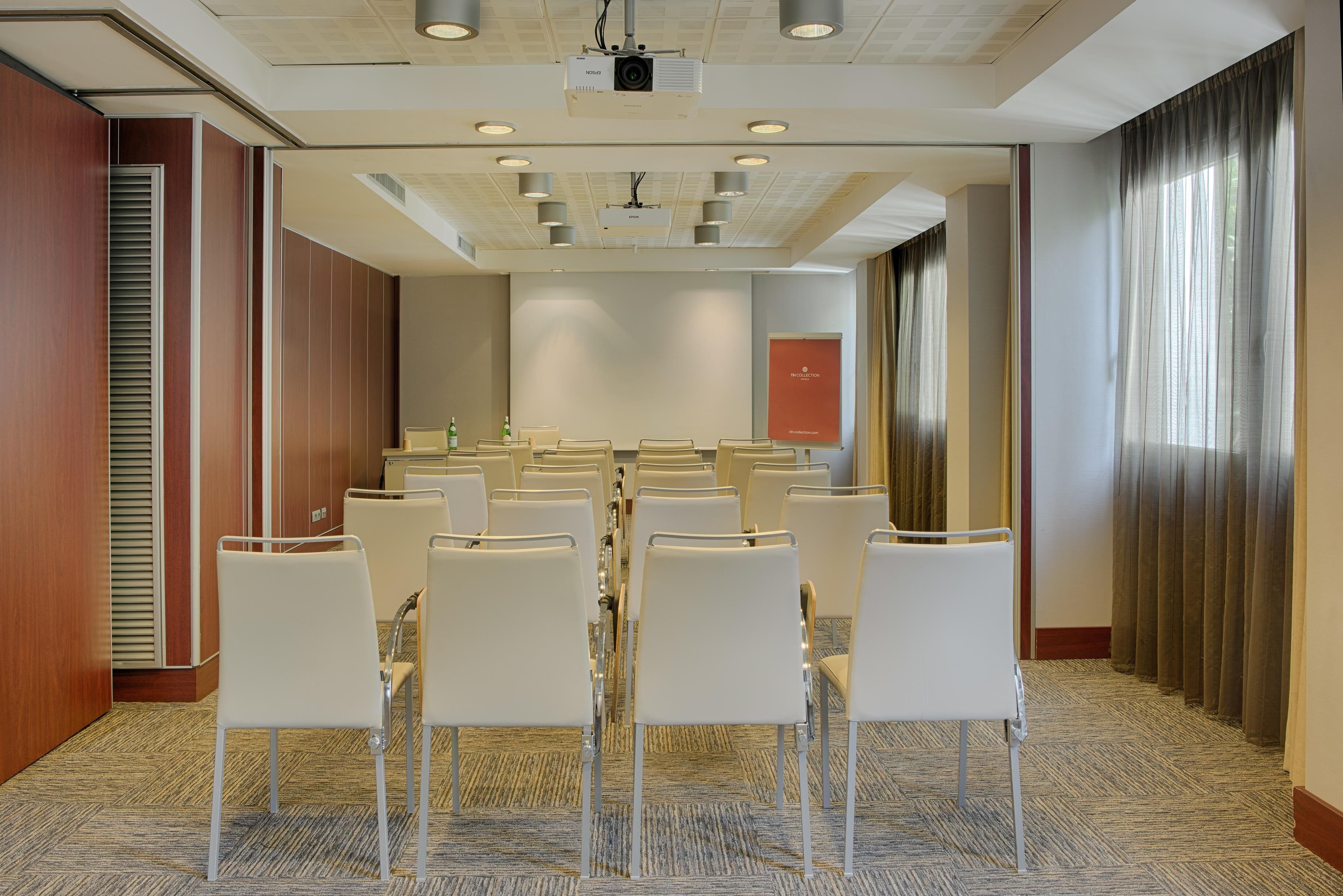 A conference room with rows of white chairs, a projector, and a red sign on the wall.
