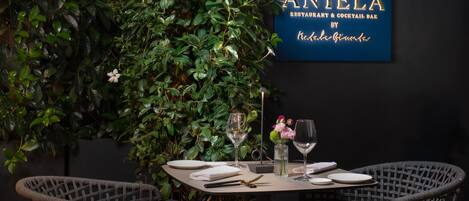 A dining table set with plates, glasses, and cutlery, surrounded by wicker chairs, with a lush green plant wall and a sign for 'ANTELA RESTAURANT & COCKTAIL BAR'.