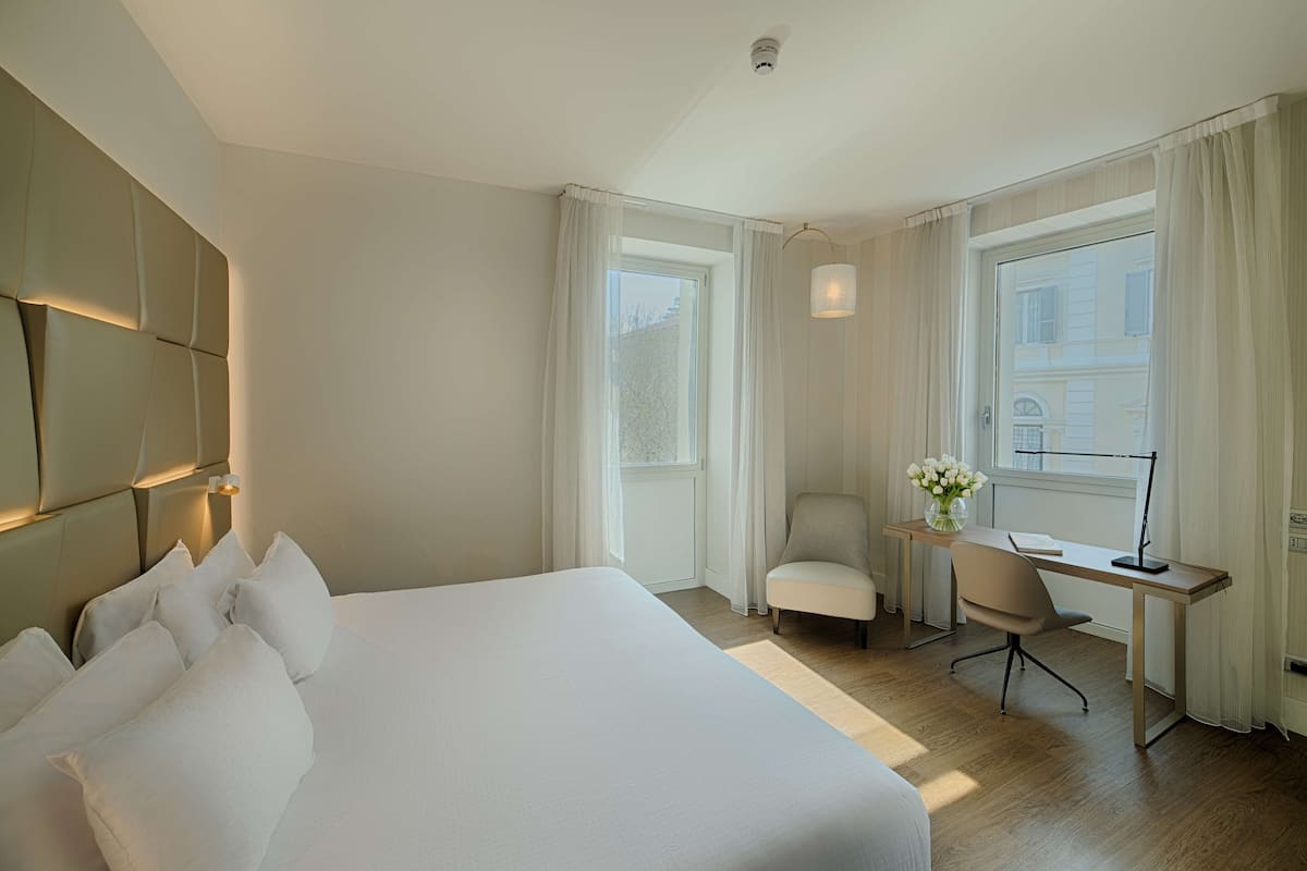 A hotel room with a large bed, a desk with a chair, and a window with curtains.