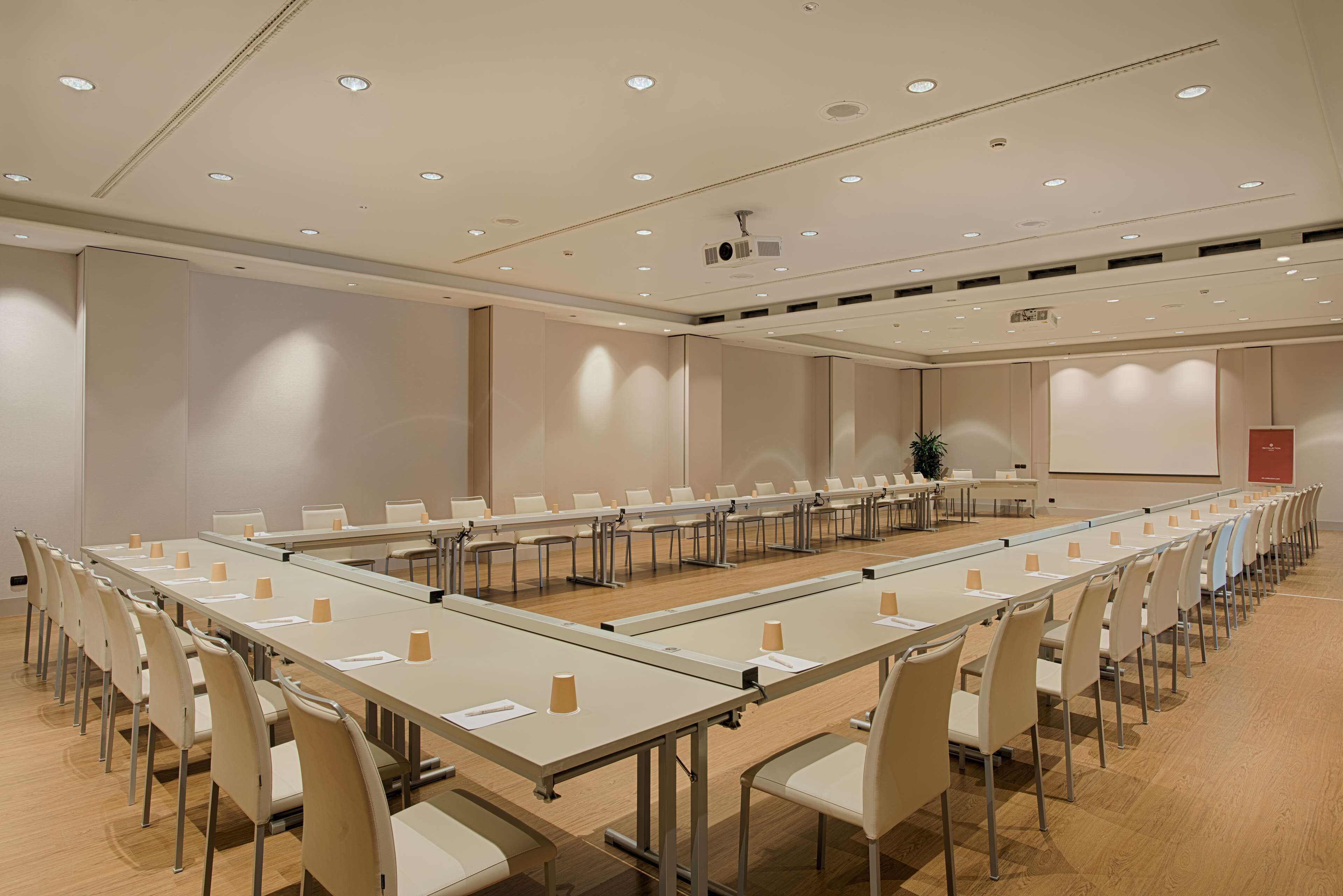 Un salón de conferencias con mesas largas, sillas y una pantalla de proyector.
