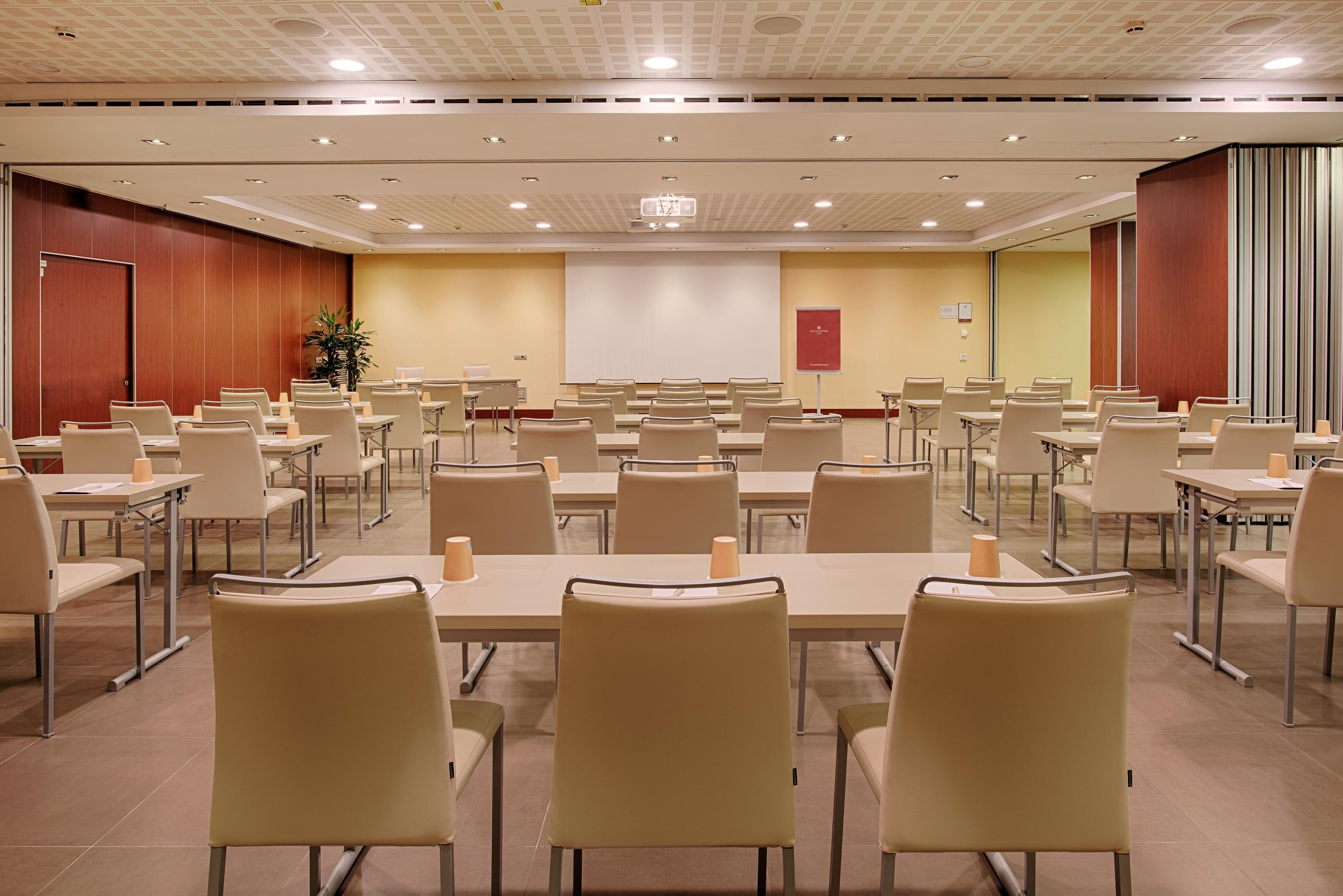 A conference room with multiple tables and chairs, a projector screen, and a large plant.