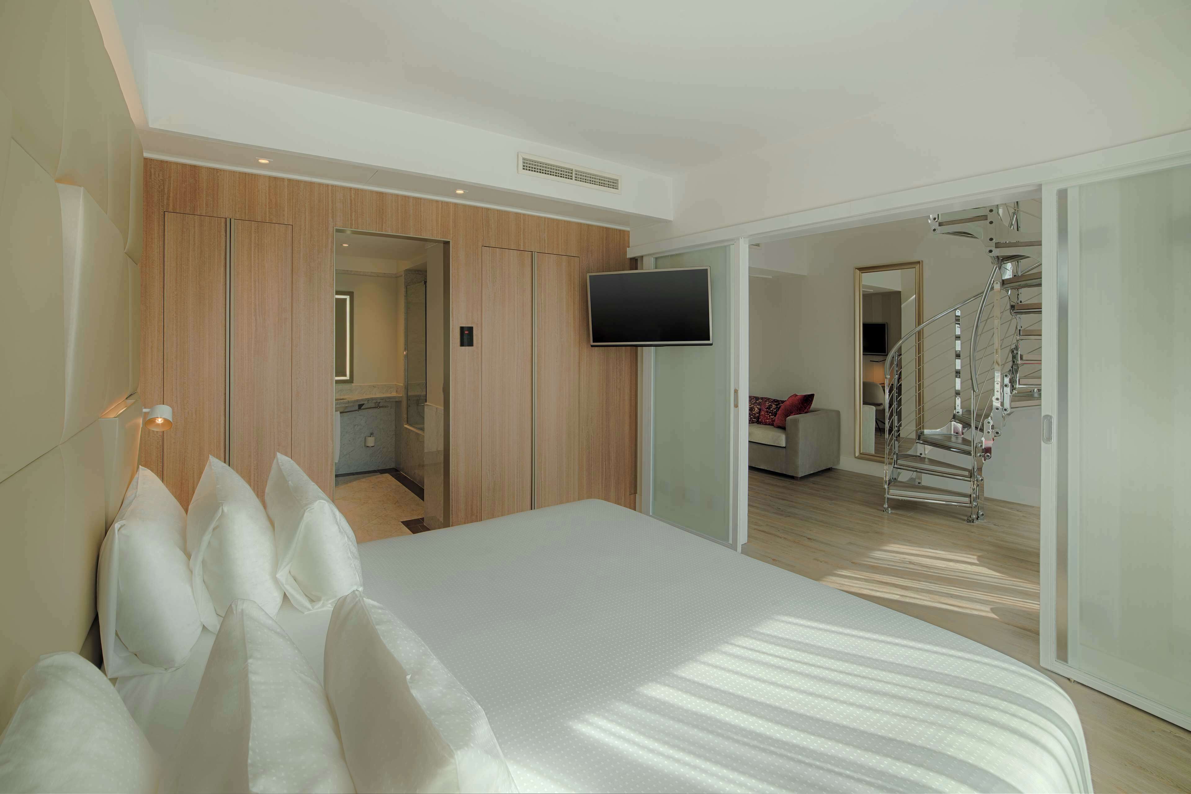 A modern hotel room with a large bed, a TV mounted on the wall, and a staircase leading to an upper floor.