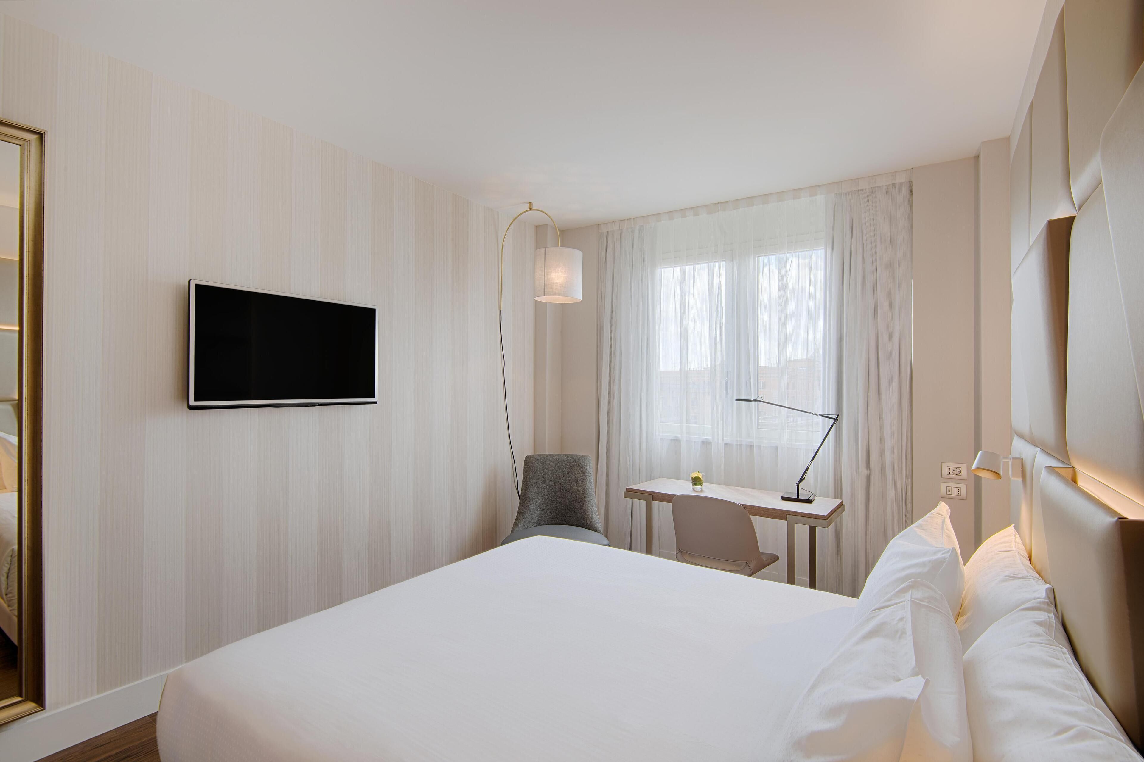 Habitación de hotel con una cama grande, un escritorio, una silla, un televisor y un ventanal con cortinas.