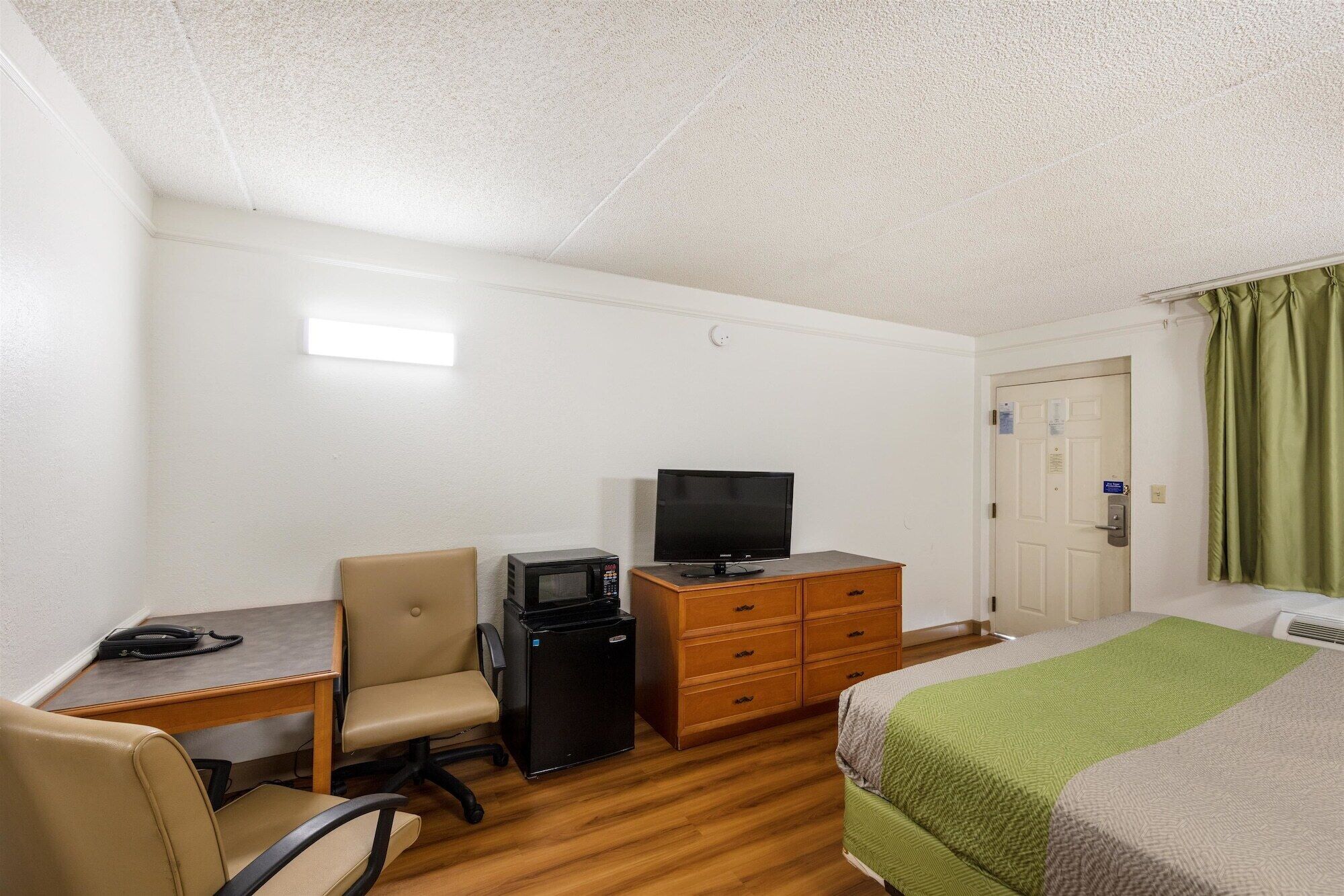 Una habitación de hotel con escritorio, silla, televisor, cómoda y cama con manta verde.