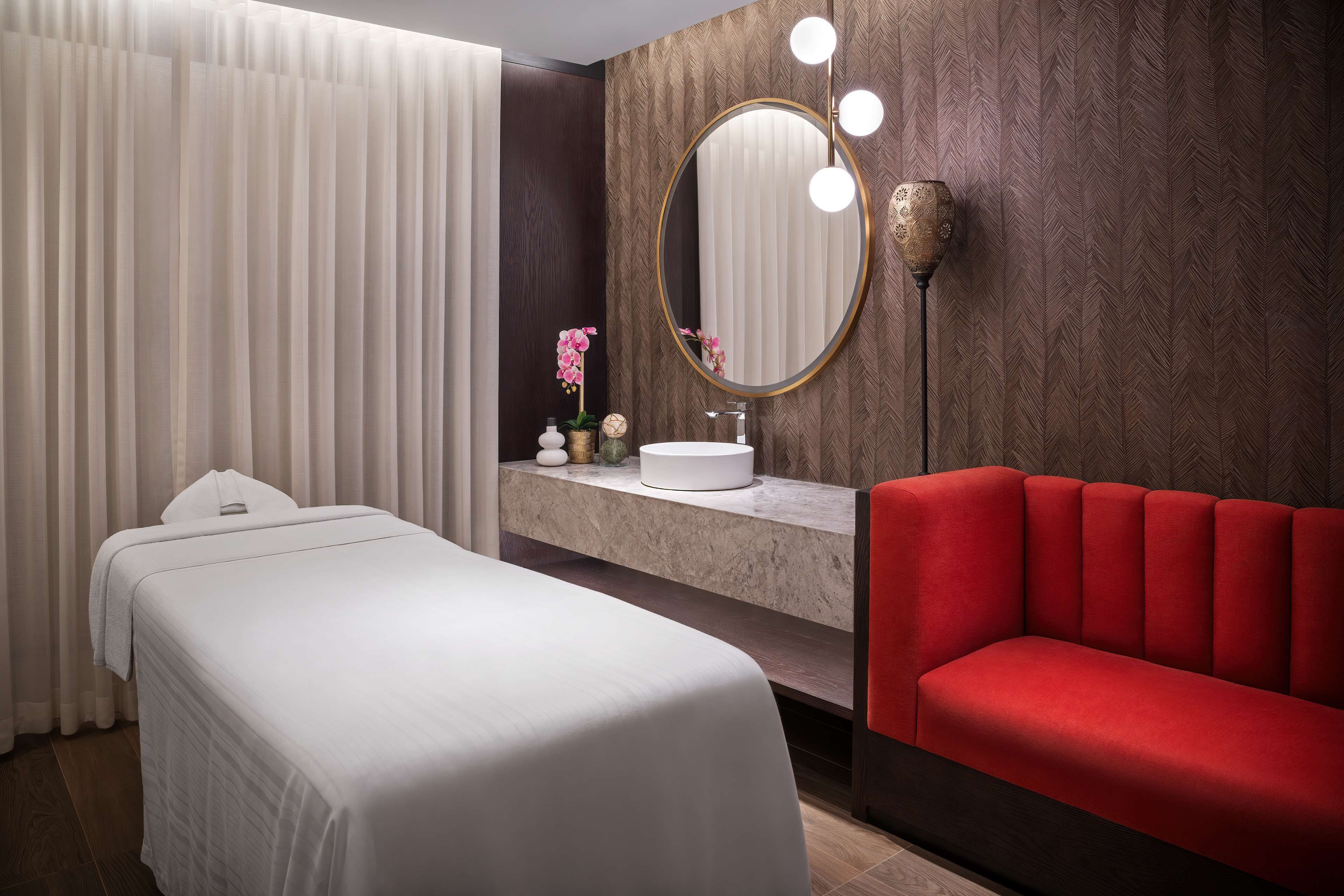 A hotel room with a bed, a red sofa, a vanity with a mirror and lights, and a potted orchid.