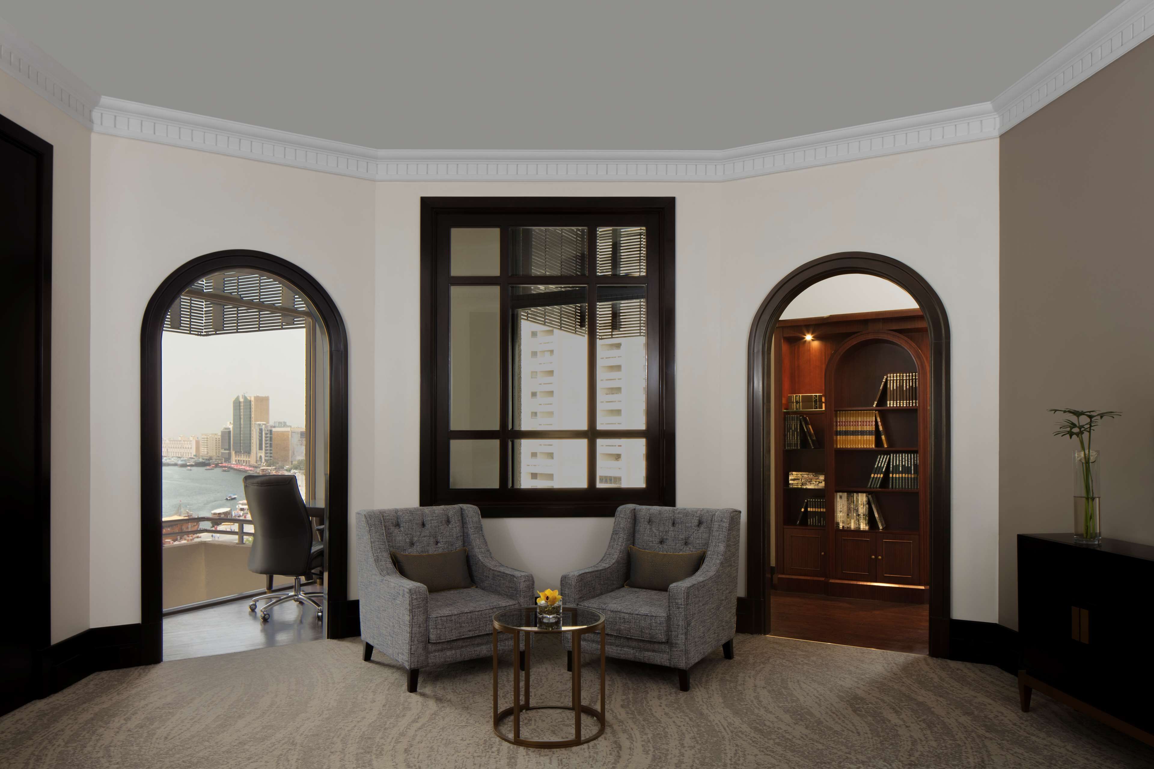 A corner room with two grey armchairs, a small round table with a vase, a bookshelf, and a large window with a city view.