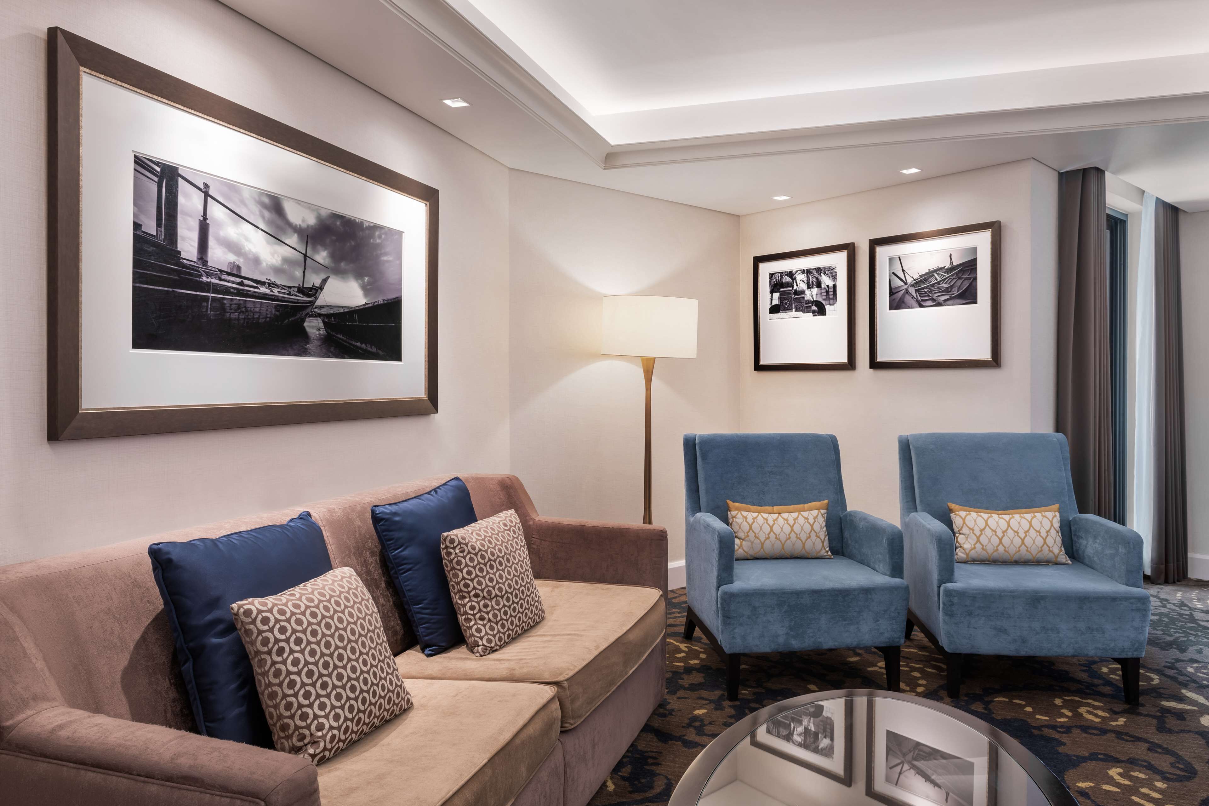 Une chambre d’hôtel avec un canapé beige, deux fauteuils bleus, une lampe sur pied et des tableaux encadrés accrochés aux murs.