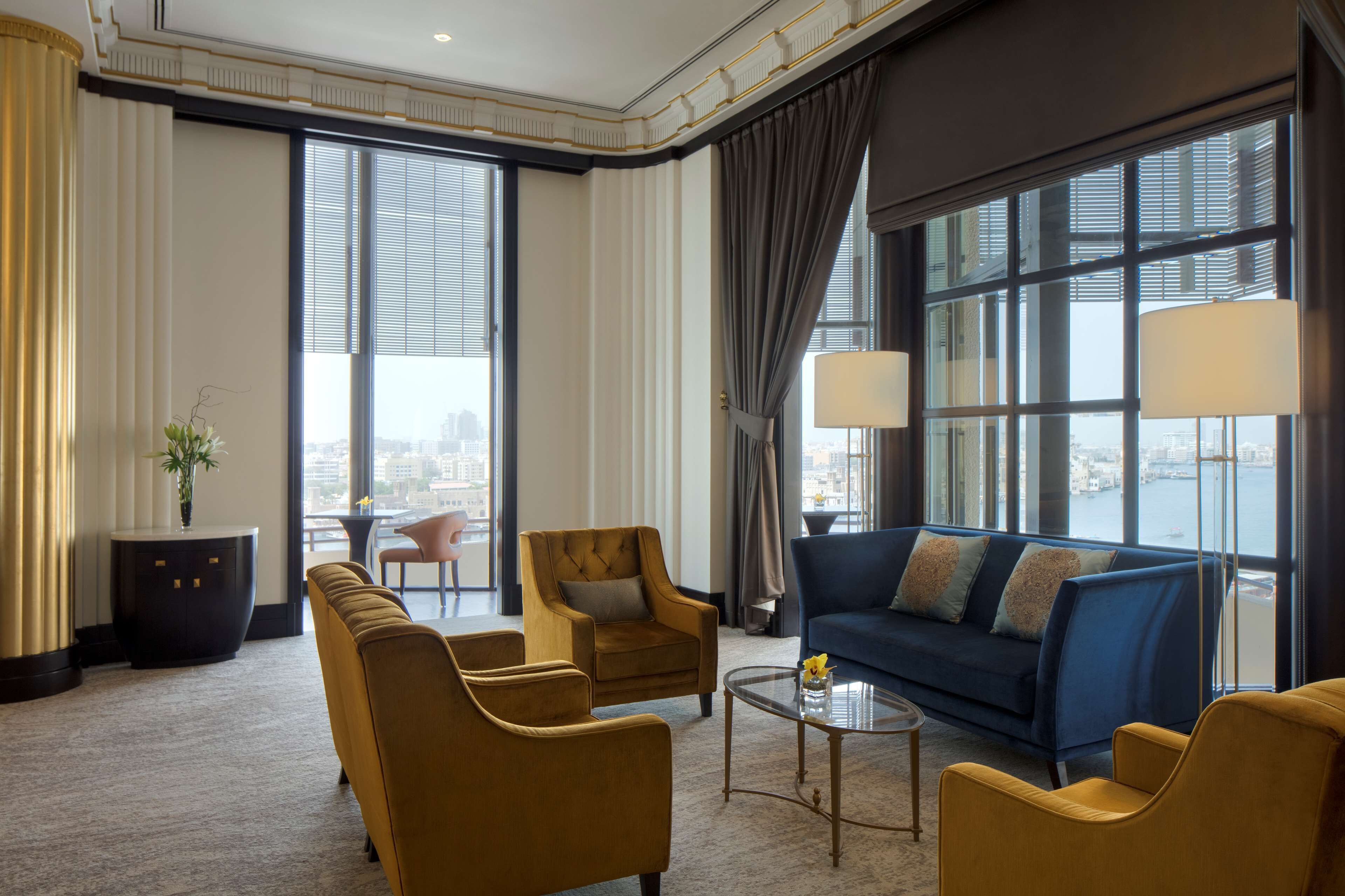 A spacious hotel room with a large window, a blue sofa, two armchairs, a coffee table, and a side table with a vase.