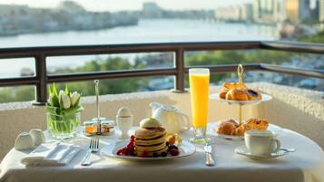 Una tavola per la colazione con pancake, croissant e un bicchiere di succo d'arancia, con vista su un fiume e sullo skyline della città.
