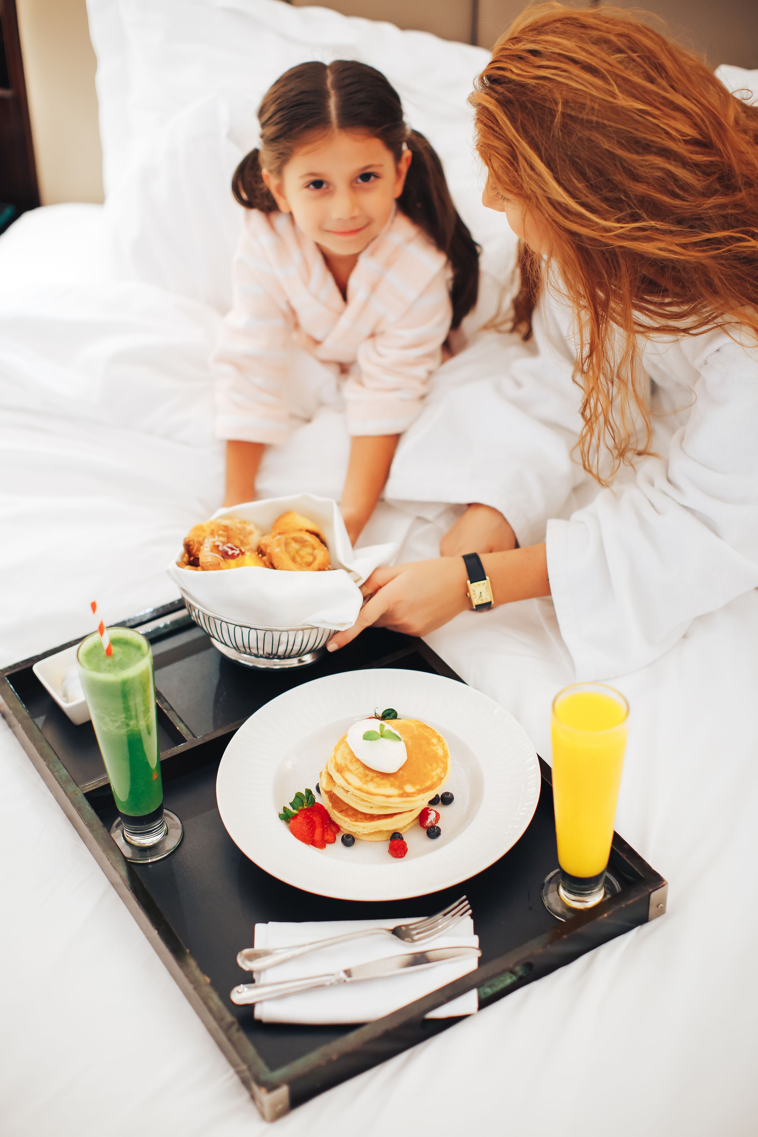 Een ontbijfwagen met pannenkoeken, een groen drankje en een glas sinaasappelsap op een bed.