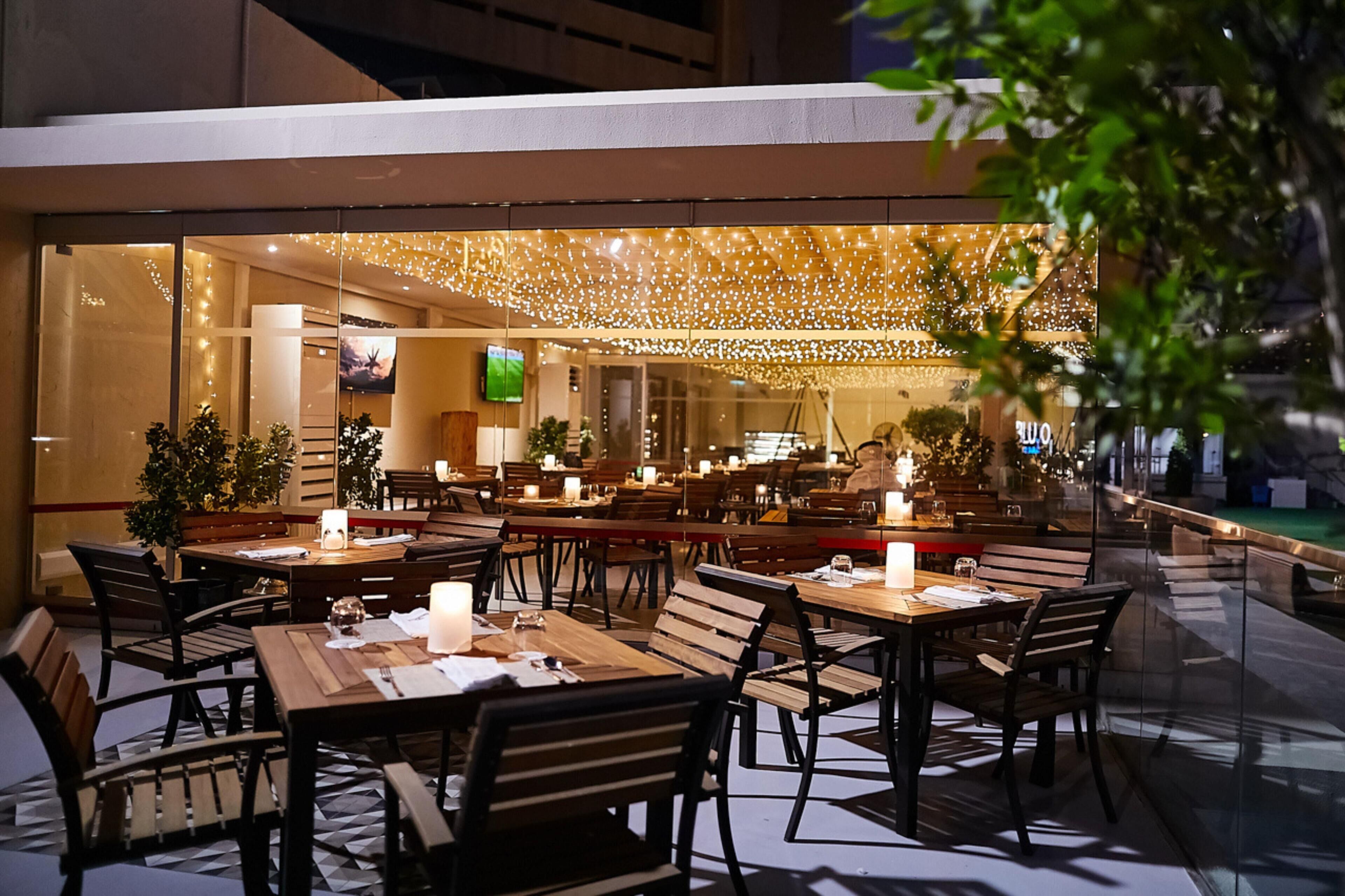 Un espace de restauration en plein air avec des tables et des chaises, des guirlandes lumineuses suspendues au-dessus et un écran de télévision visible à l’intérieur.