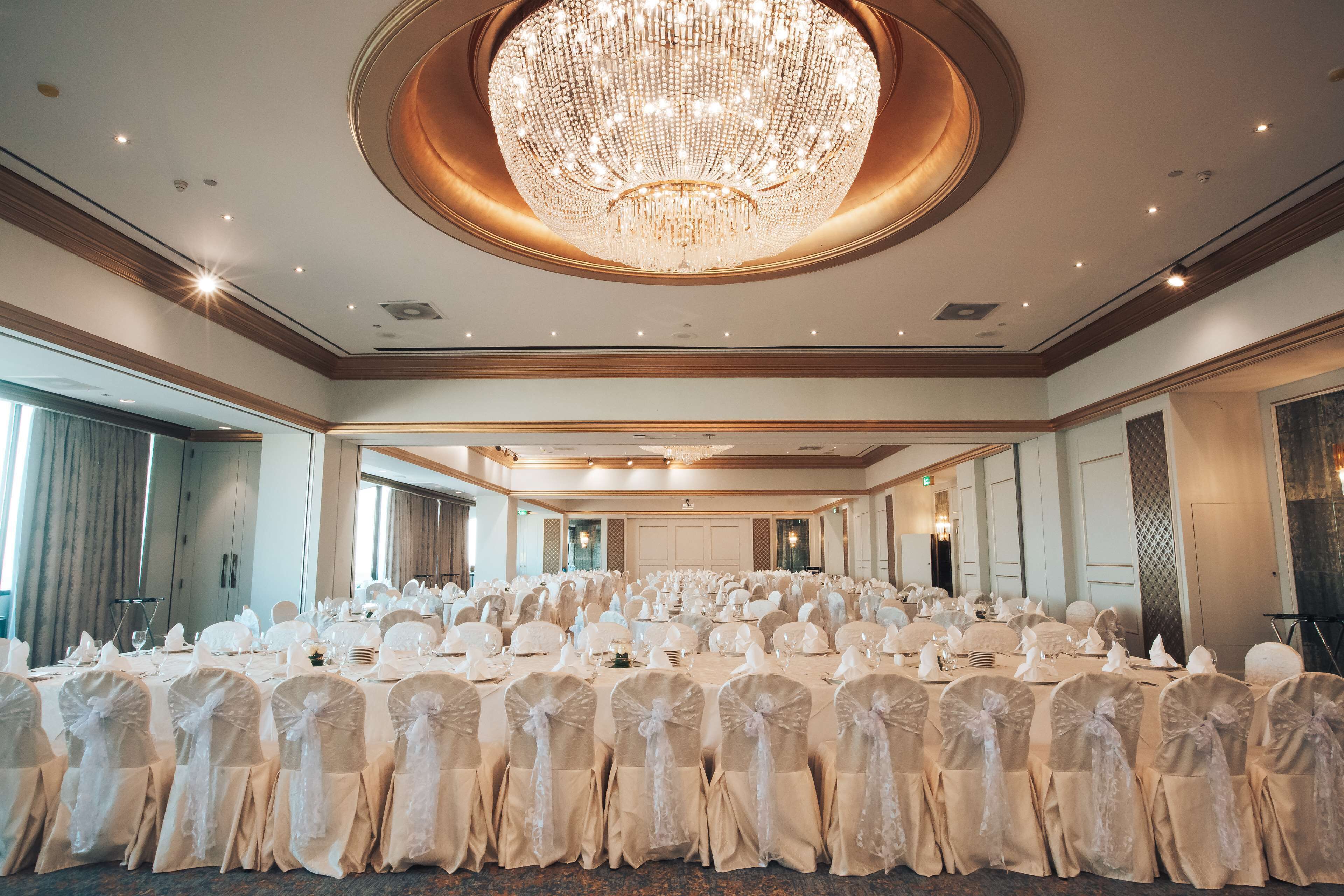 A banquet hall with rows of tables, white chairs, and a large chandelier.