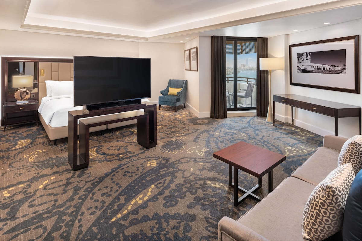 A hotel room with a large bed, a flat-screen TV, a sofa, a small table, a chair, a desk, and a balcony with a view.
