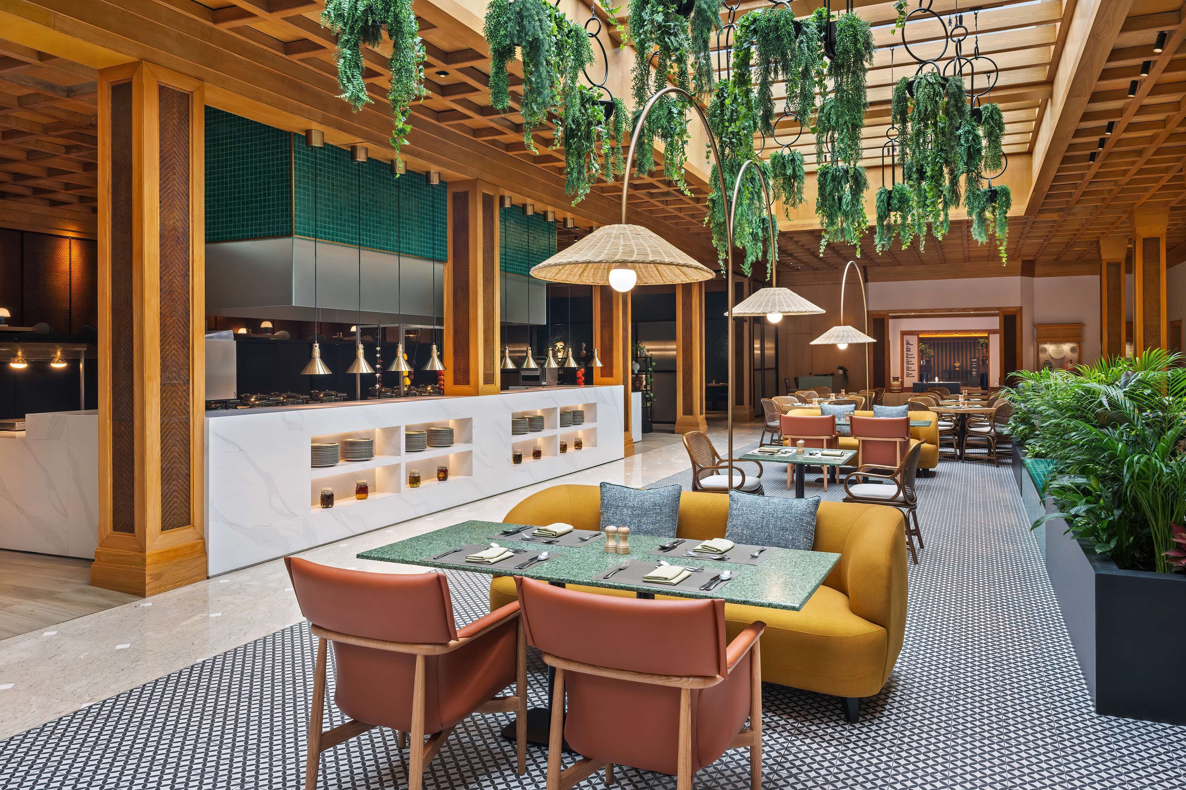 A modern restaurant with a central dining area featuring yellow sofas and orange chairs, a kitchen in the background, and hanging plants.