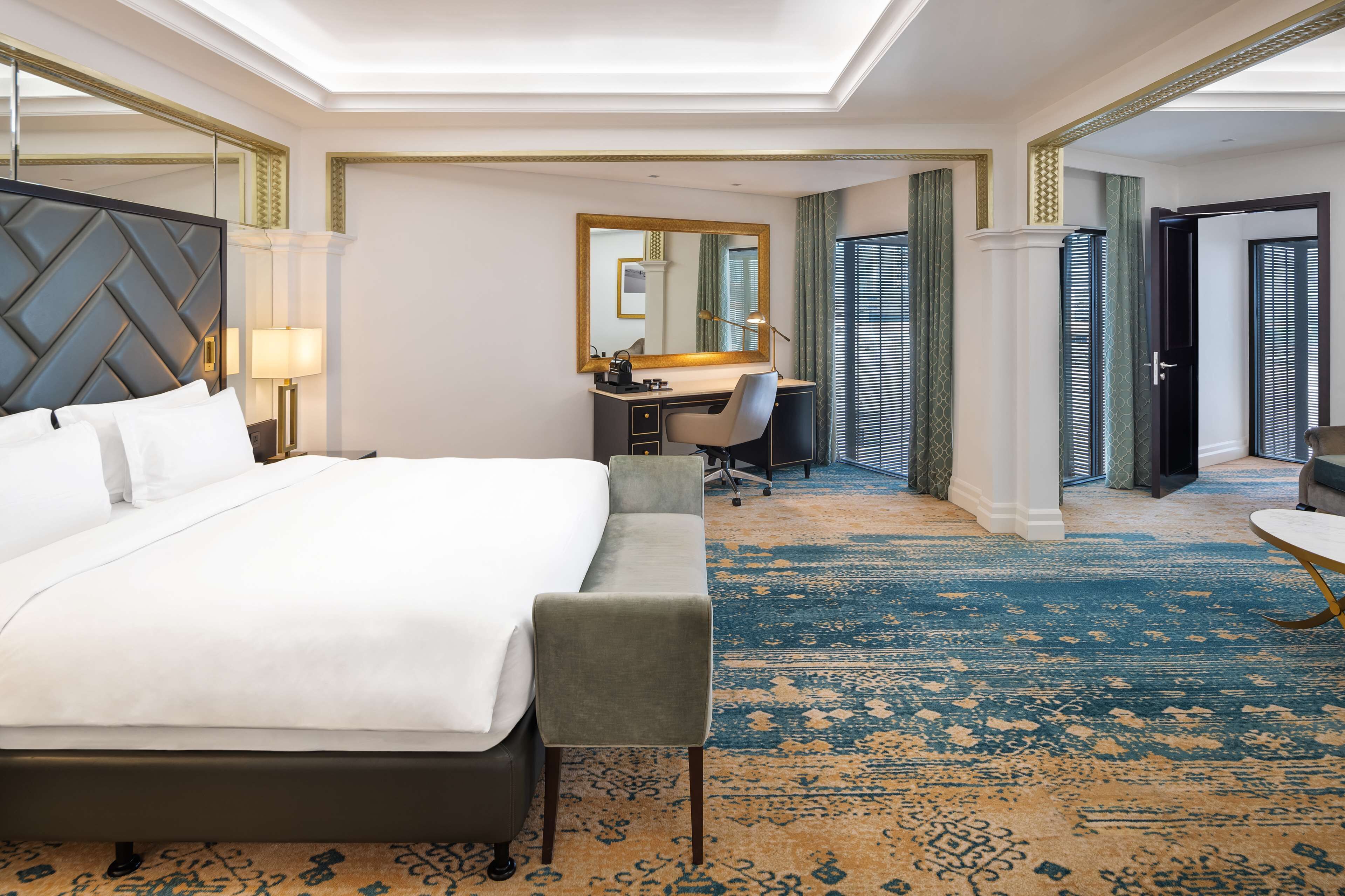 A hotel room with a large bed, a desk with a chair, a mirror, and a carpeted floor.